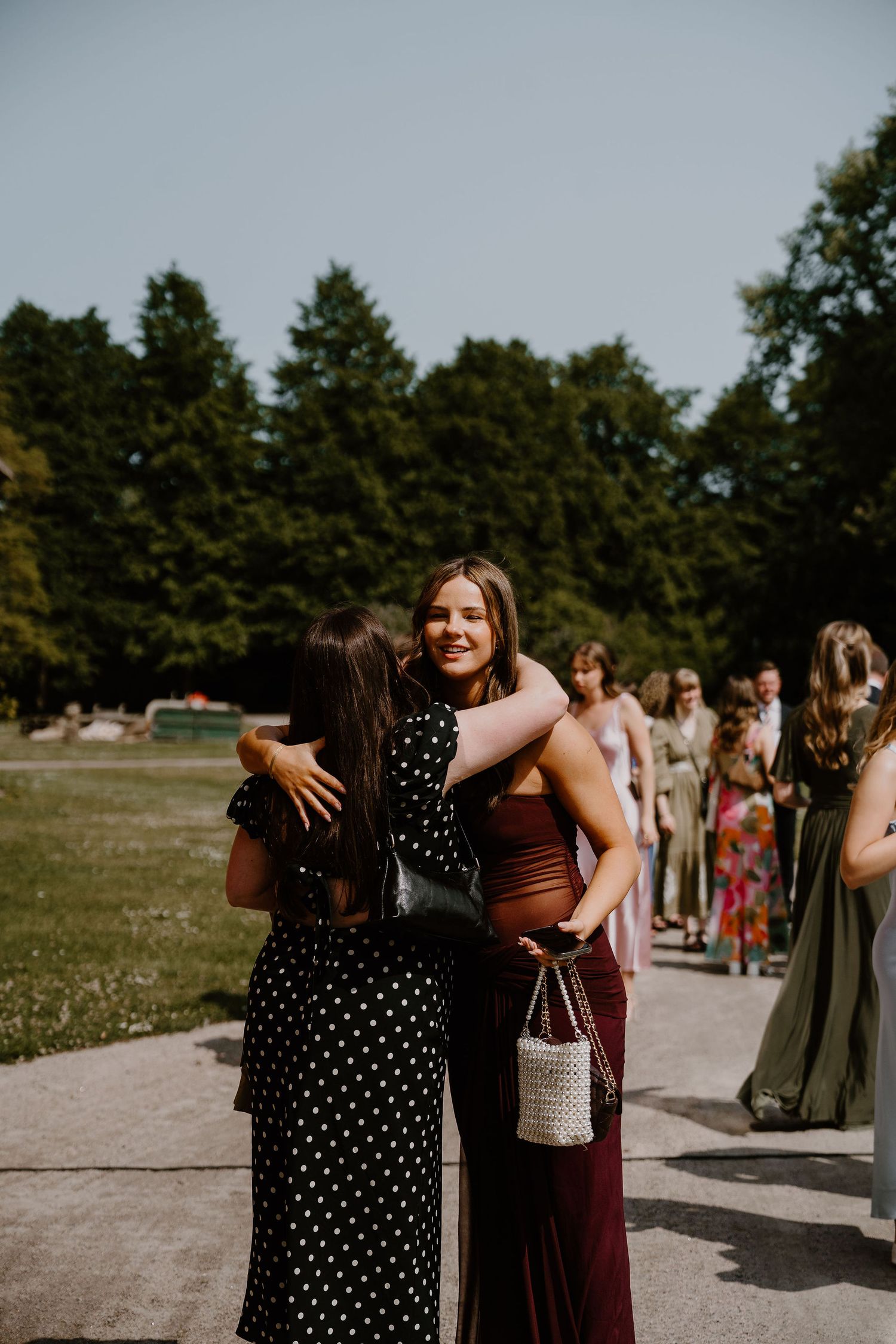 Wedding guests in groups gather outdoors on a sunny day with green grass and trees in the background.