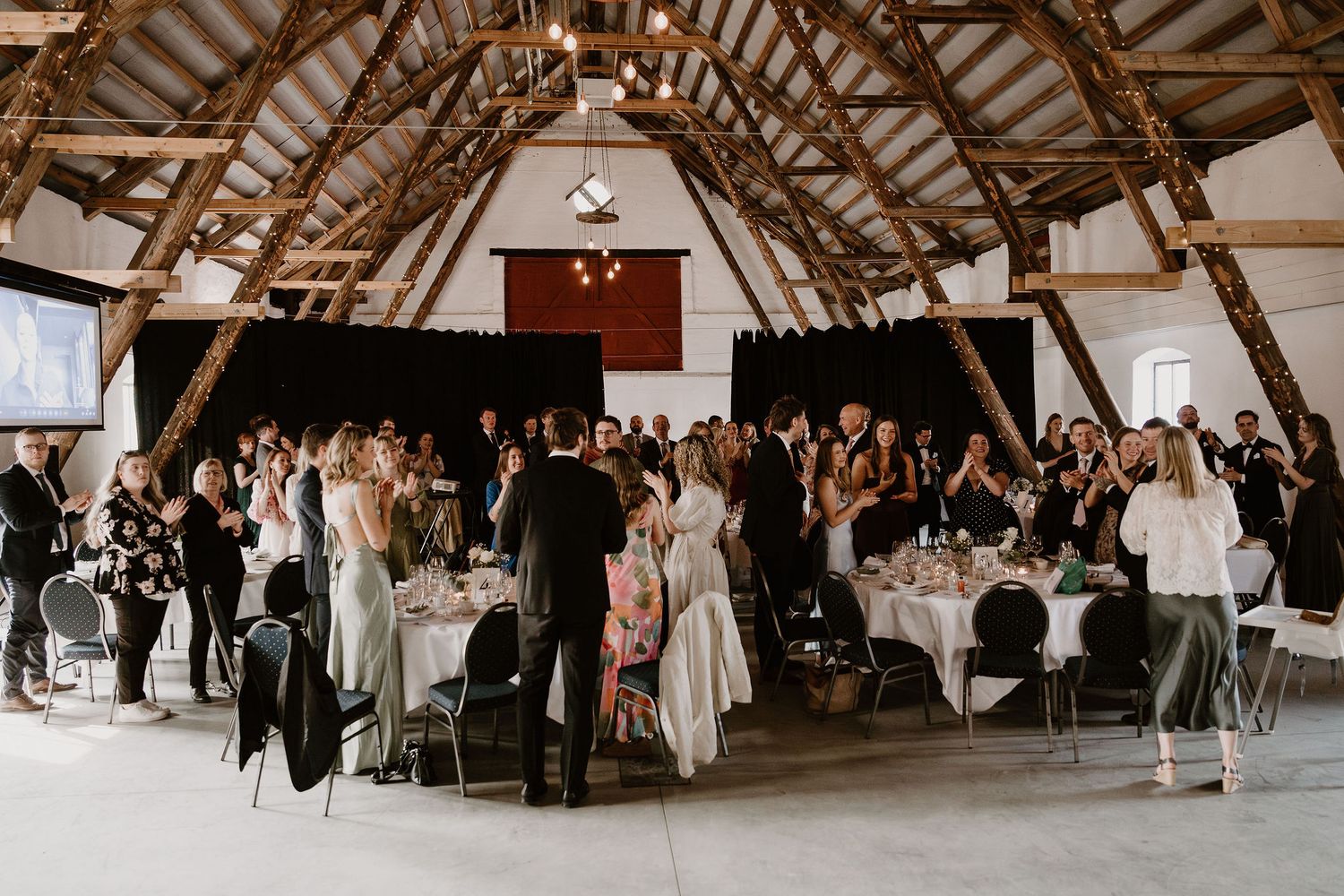 Wedding guests mingle and socialize during a reception in a rustic barn venue with exposed wooden beams and warm lighting.