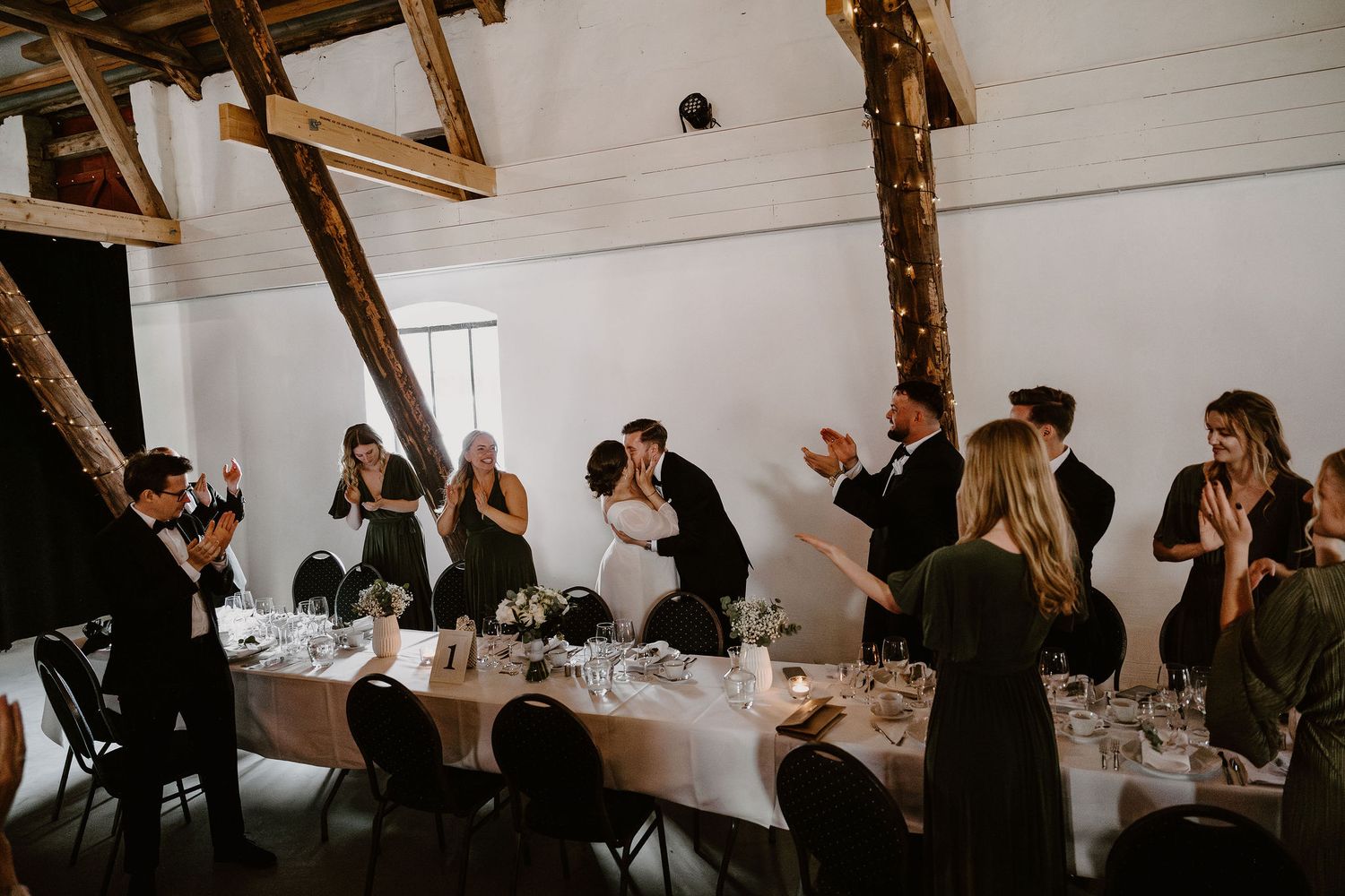 Wedding guests in formal attire gather around a long white dining table at an intimate indoor reception venue.