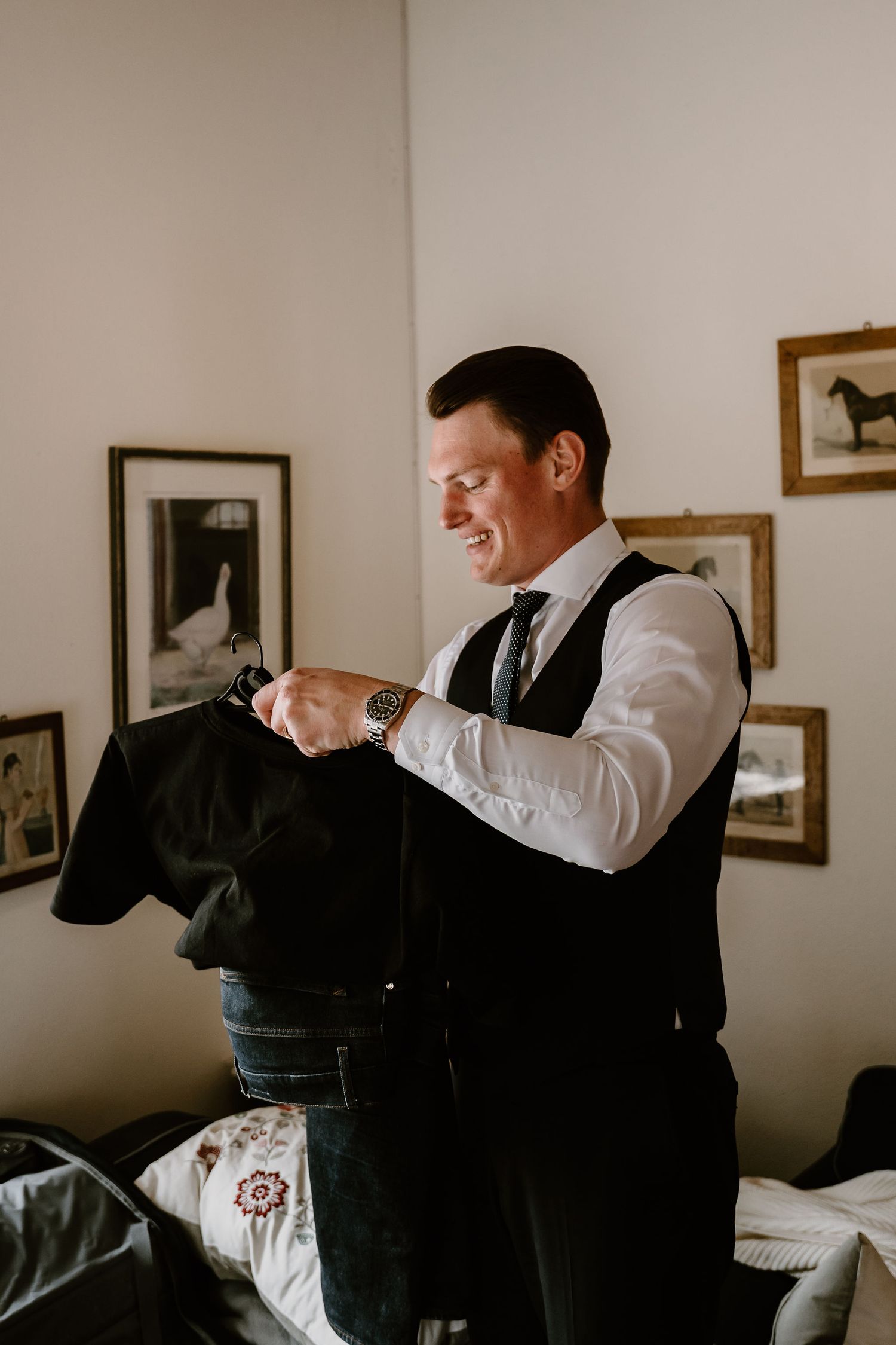 A person in formal attire with a white shirt and suspenders adjusts clothing while getting ready indoors.
