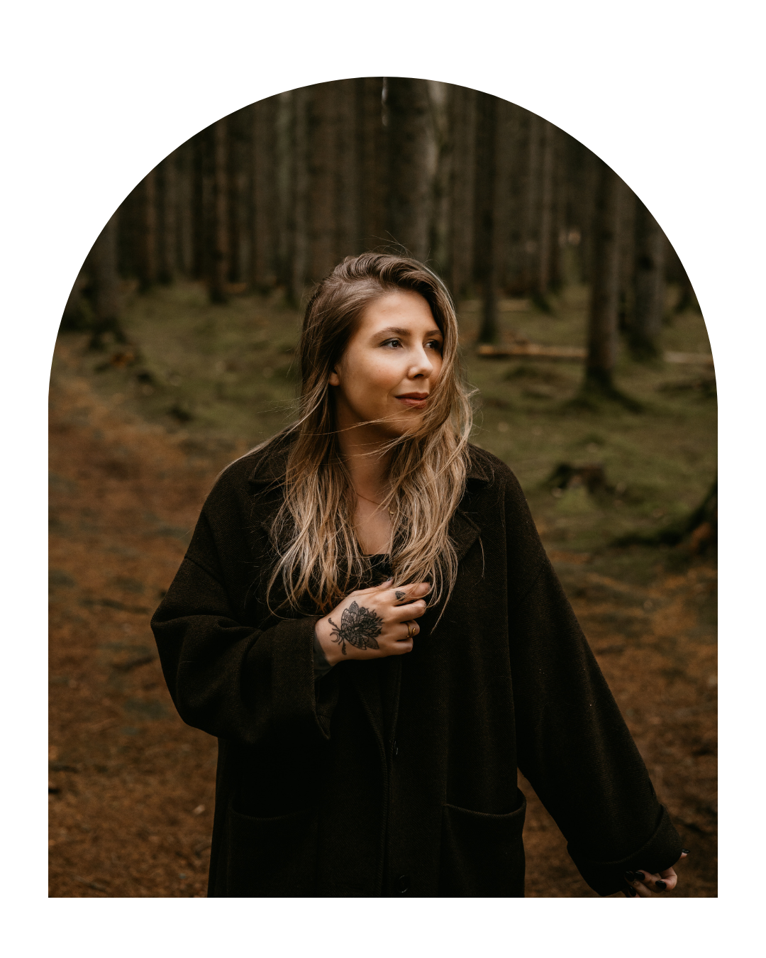 Person in a black coat stands in moody forest setting with warm earthy tones and natural lighting.