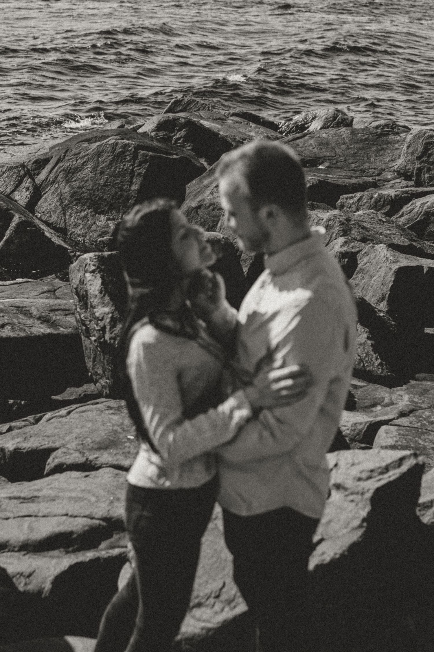 A couple stands embracing on rocky coastal shoreline in black and white photograph.