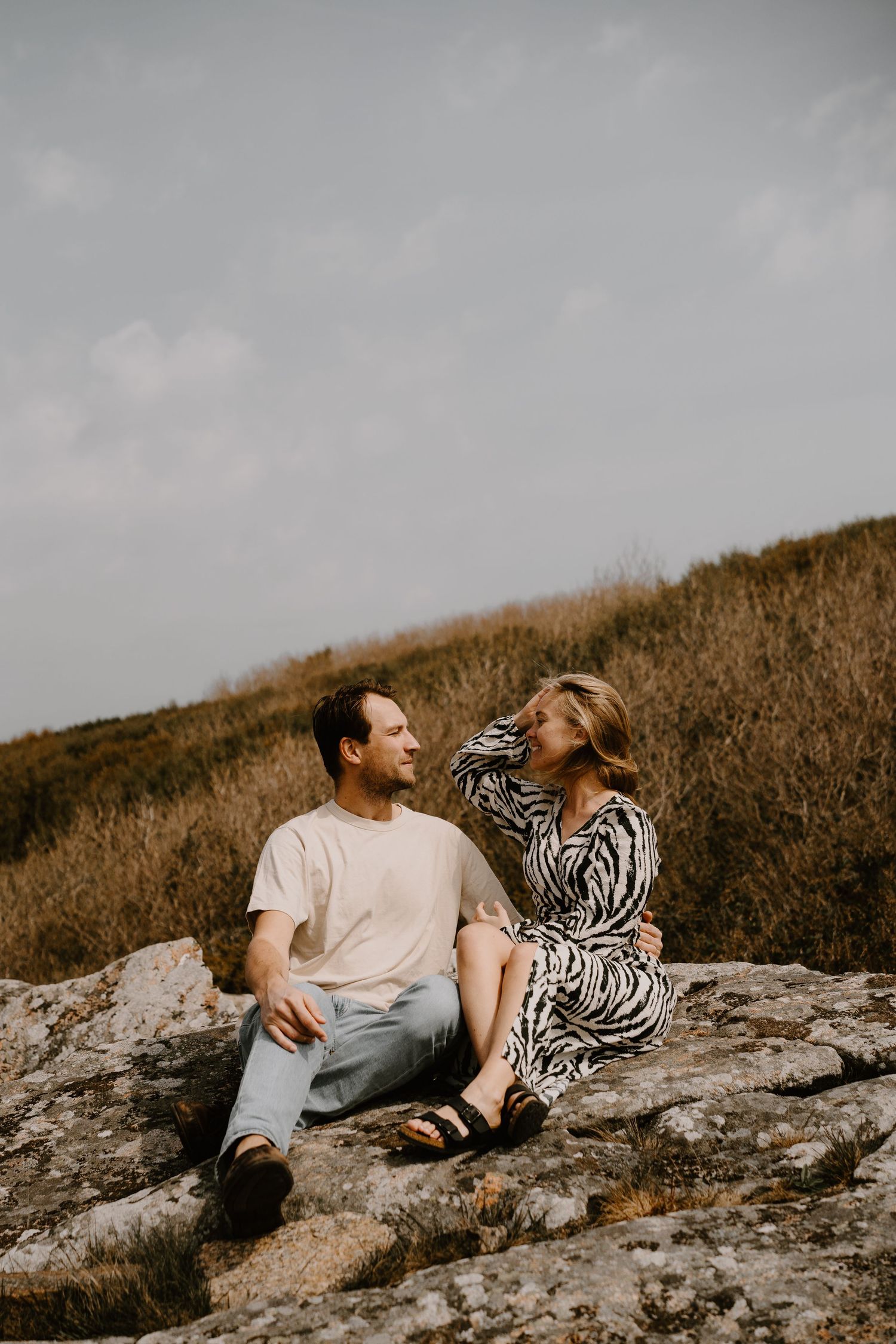 A couple laughs together while sitting on a rocky hillside during golden hour.