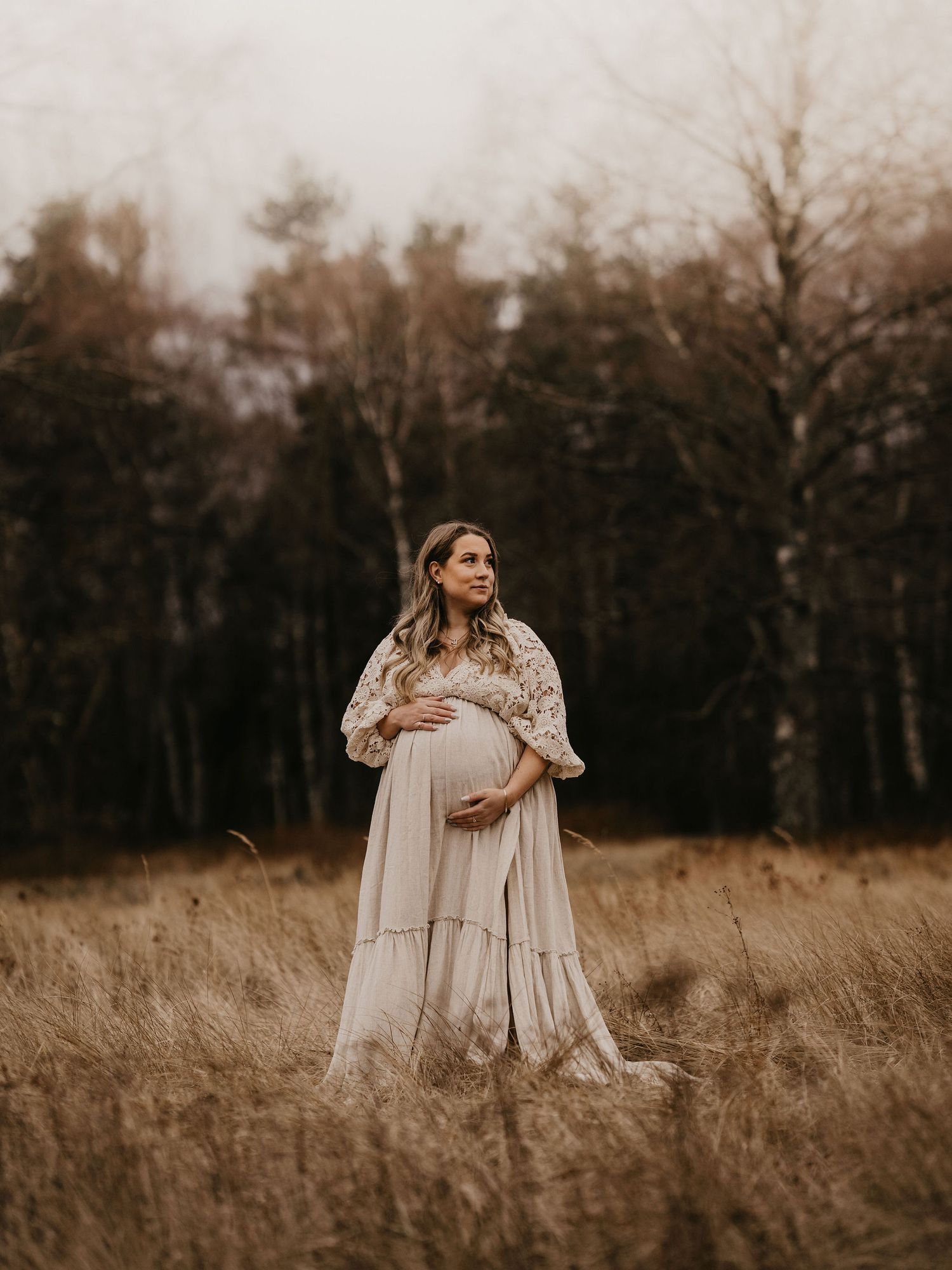 A pregnant woman in a flowing cream dress stands in a golden autumn field at dusk with bare trees in the background.