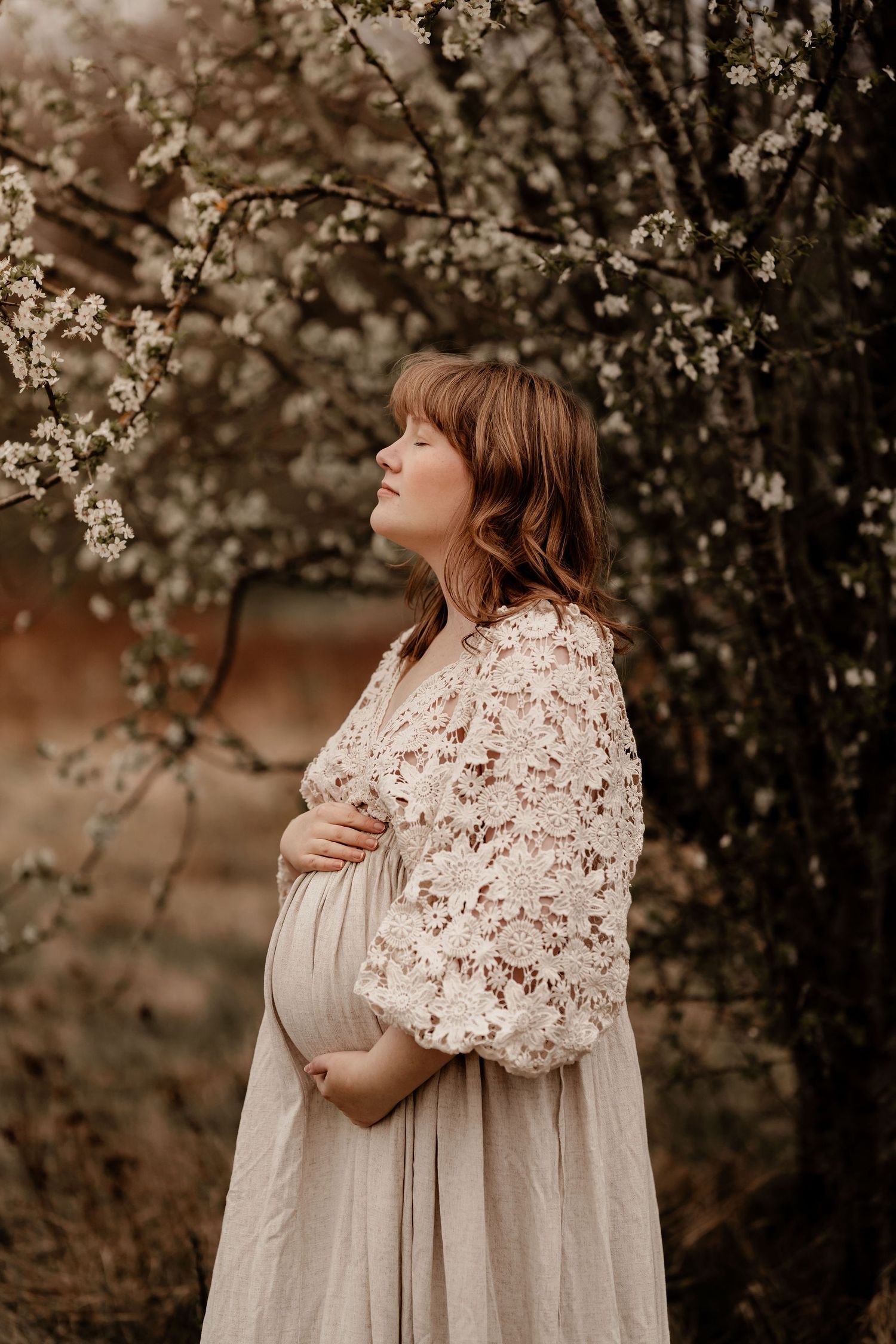 Expectant mother in floral lace dress poses serenely under blooming spring tree branches at sunset.