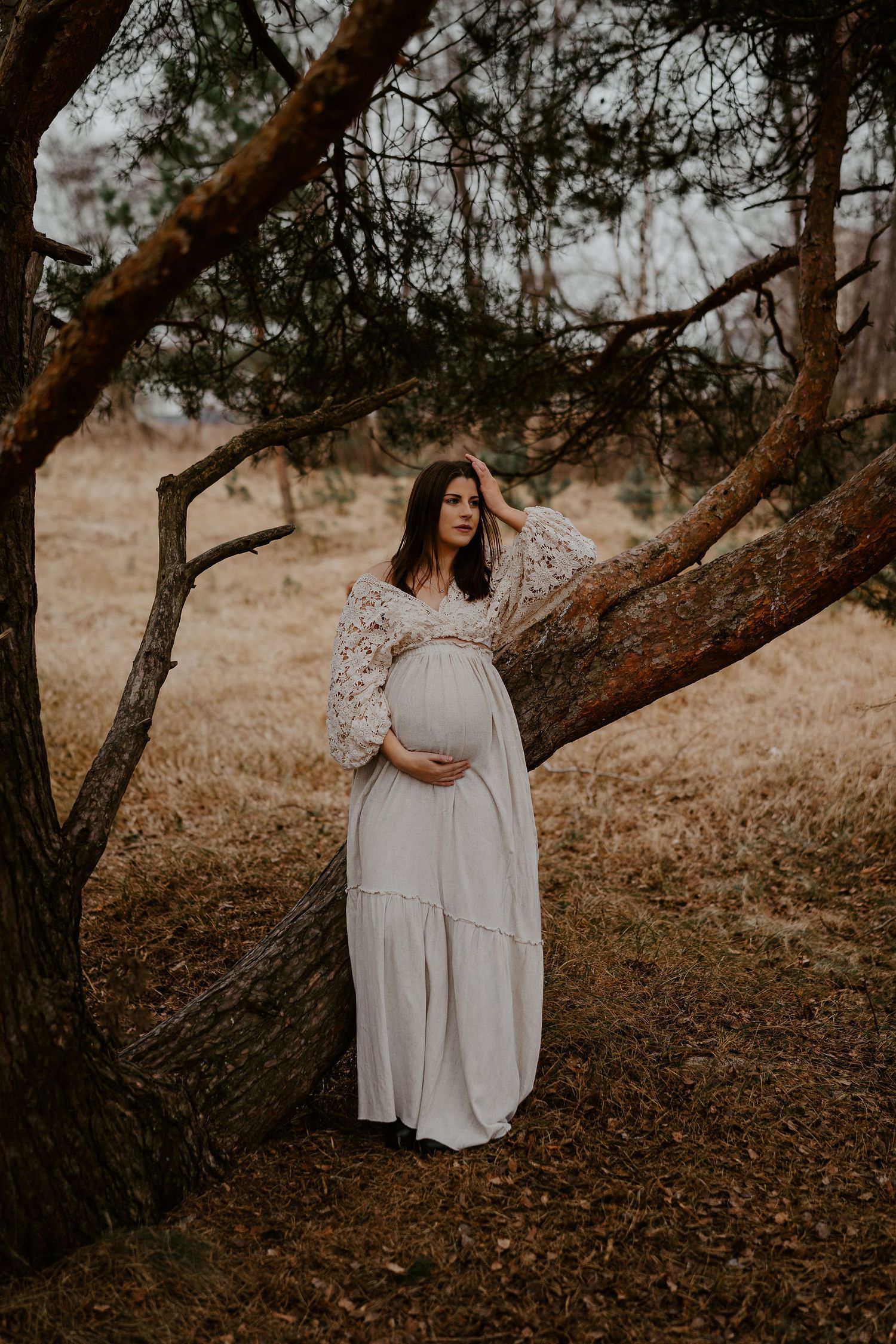 A pregnant woman in a flowing white lace dress stands beneath a large tree in a moody outdoor setting.