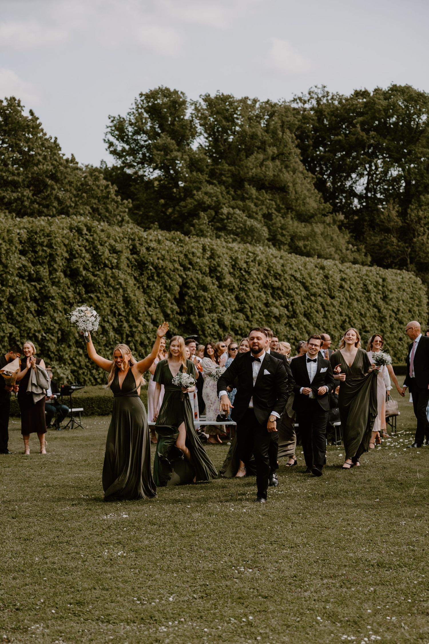 Wedding party celebrating and walking joyfully on a green lawn with tall hedges and trees in the background.