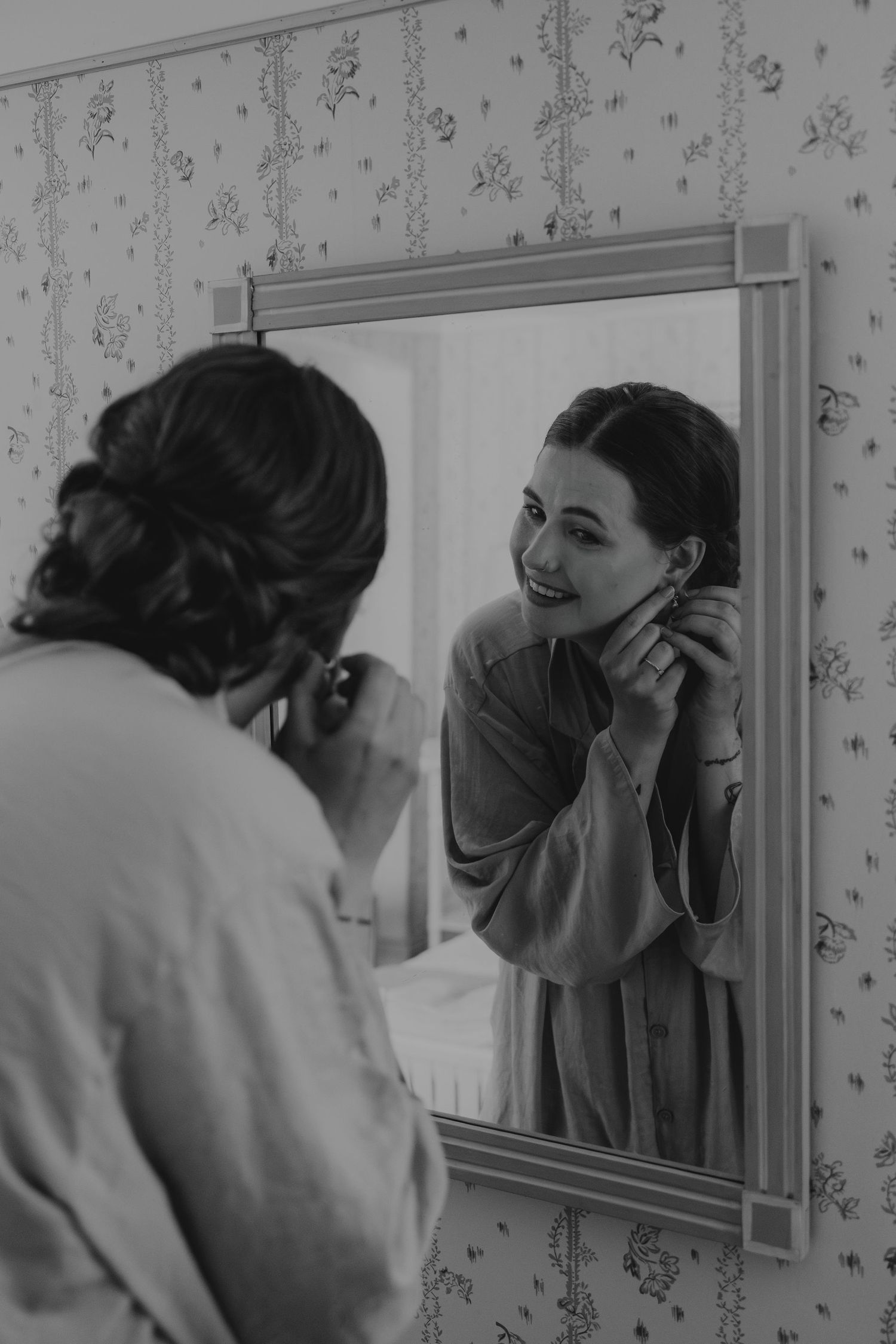 A vintage black and white photo showing a reflection in a bathroom mirror against floral wallpaper.