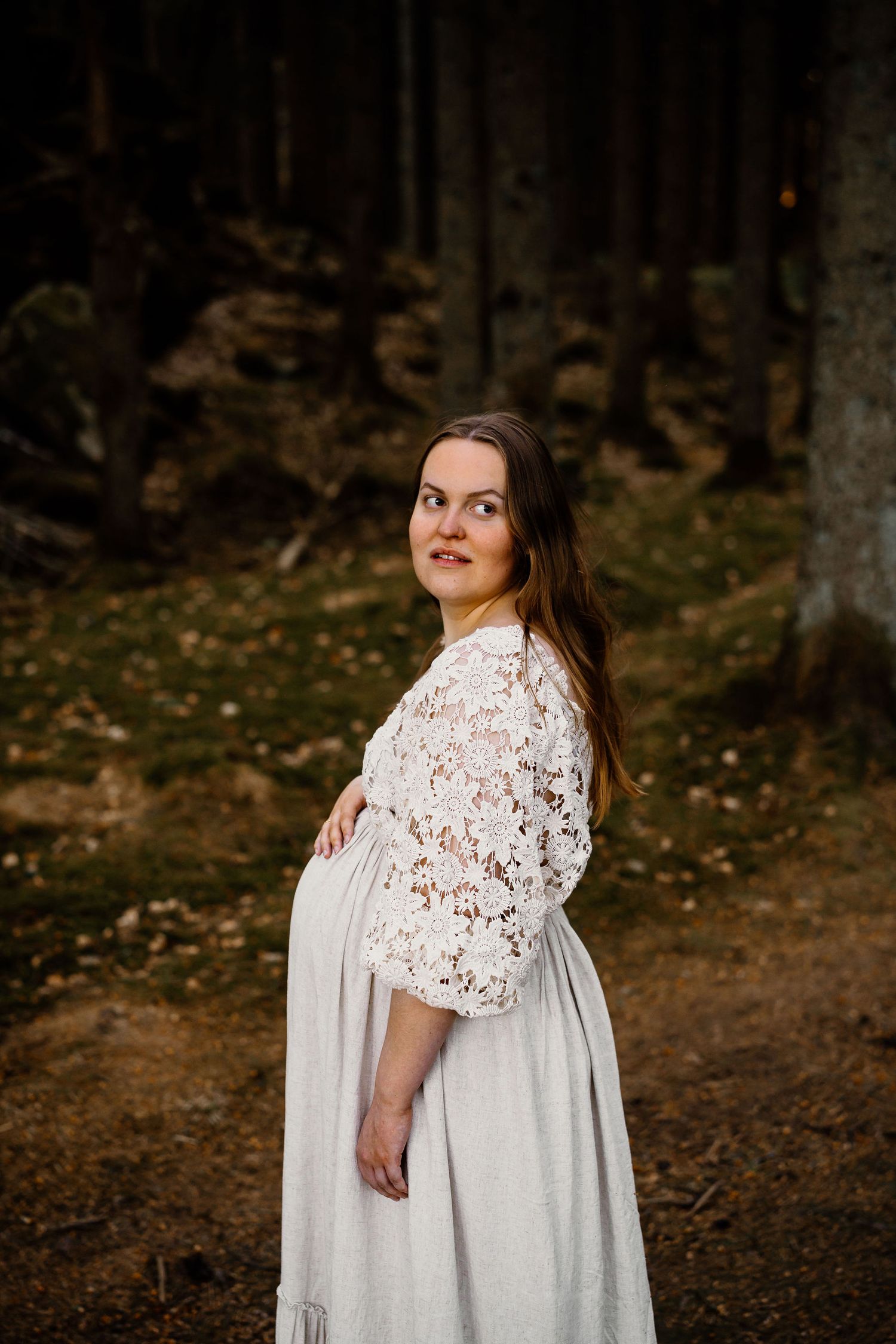 A pregnant woman stands in a forest wearing a white lace dress and looking to the side in moody autumn lighting.