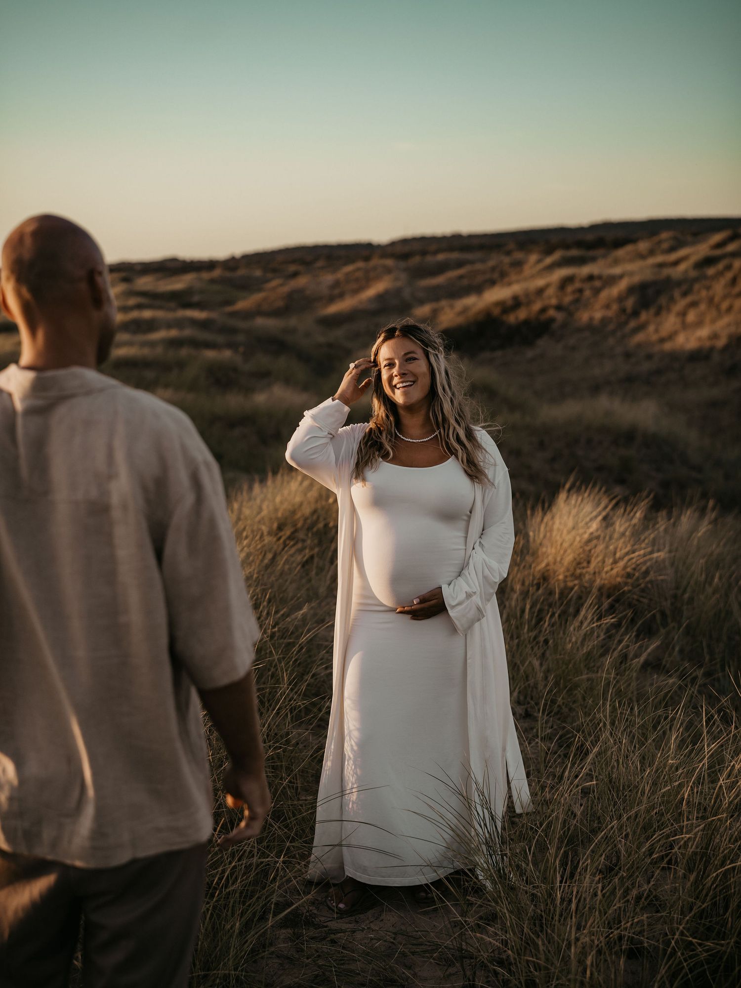 Maternity photo shoot at sunset in a grassy field with golden light illuminating a flowing white dress and cardigan.