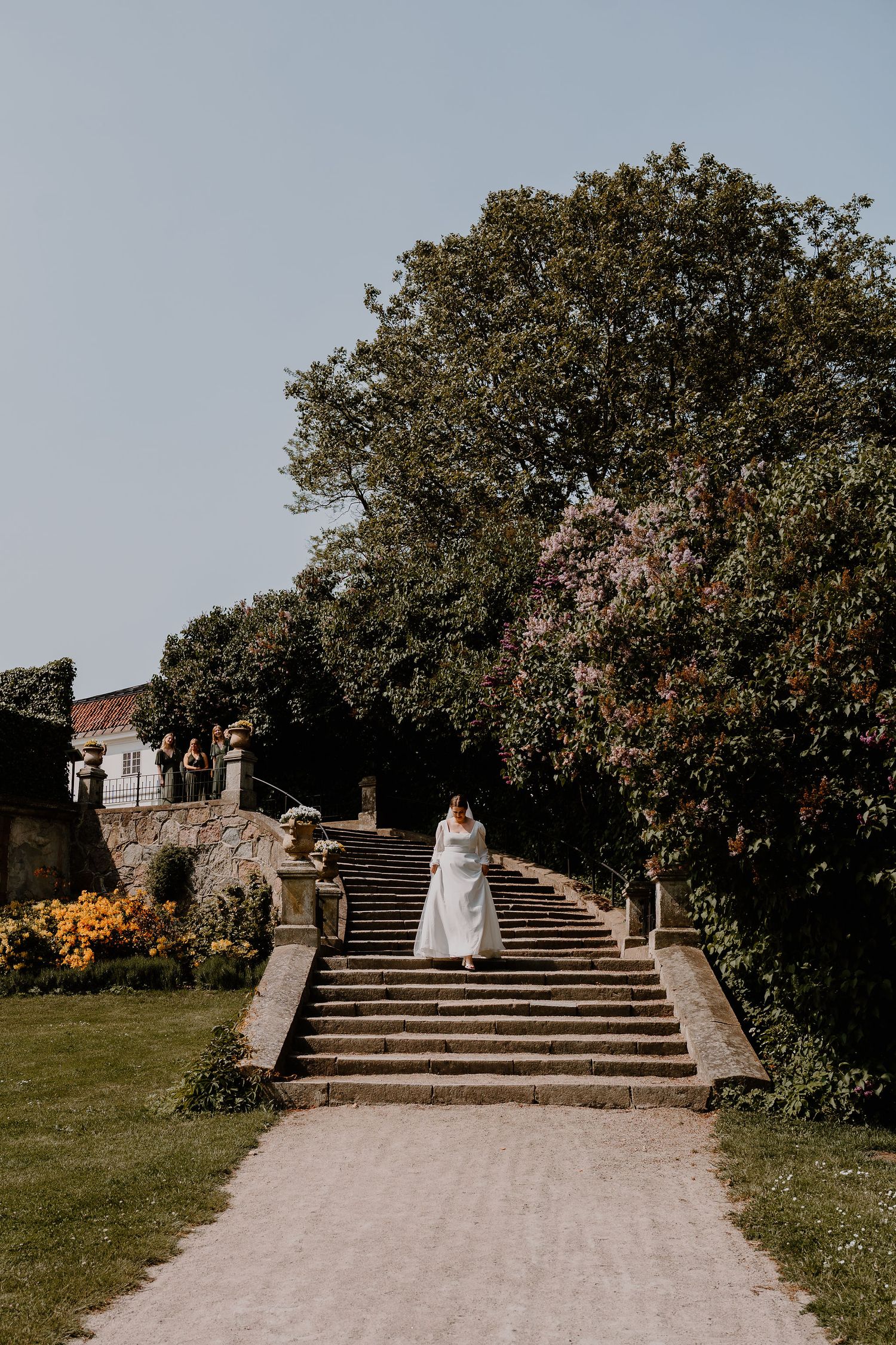 A wedding couple walks up grand stone steps surrounded by lush trees and gardens at a historic estate.