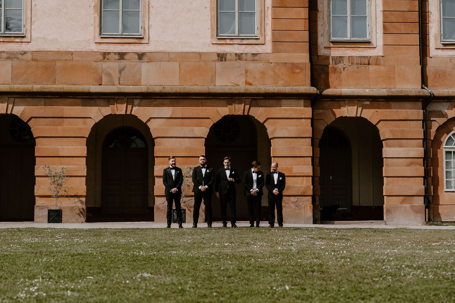 Group in formal black attire stands against arched colonnade of historic terracotta building on grassy lawn.