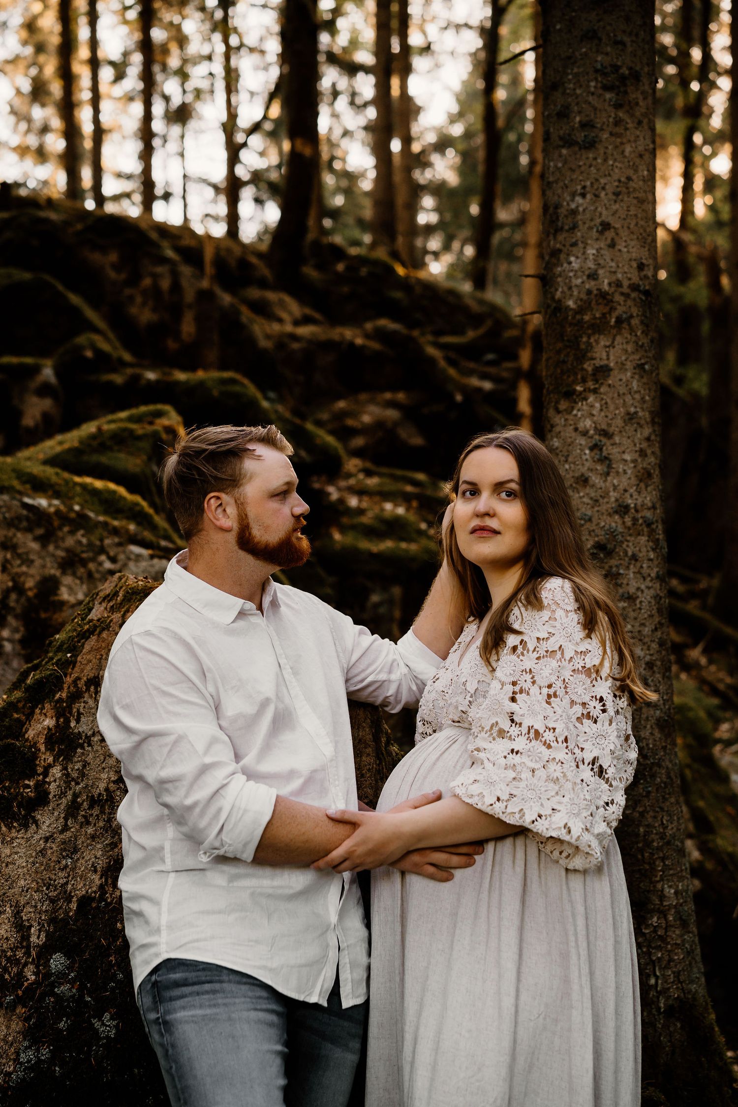A couple shares an intimate moment in front of mossy rocks and towering trees during a sunset forest photoshoot.