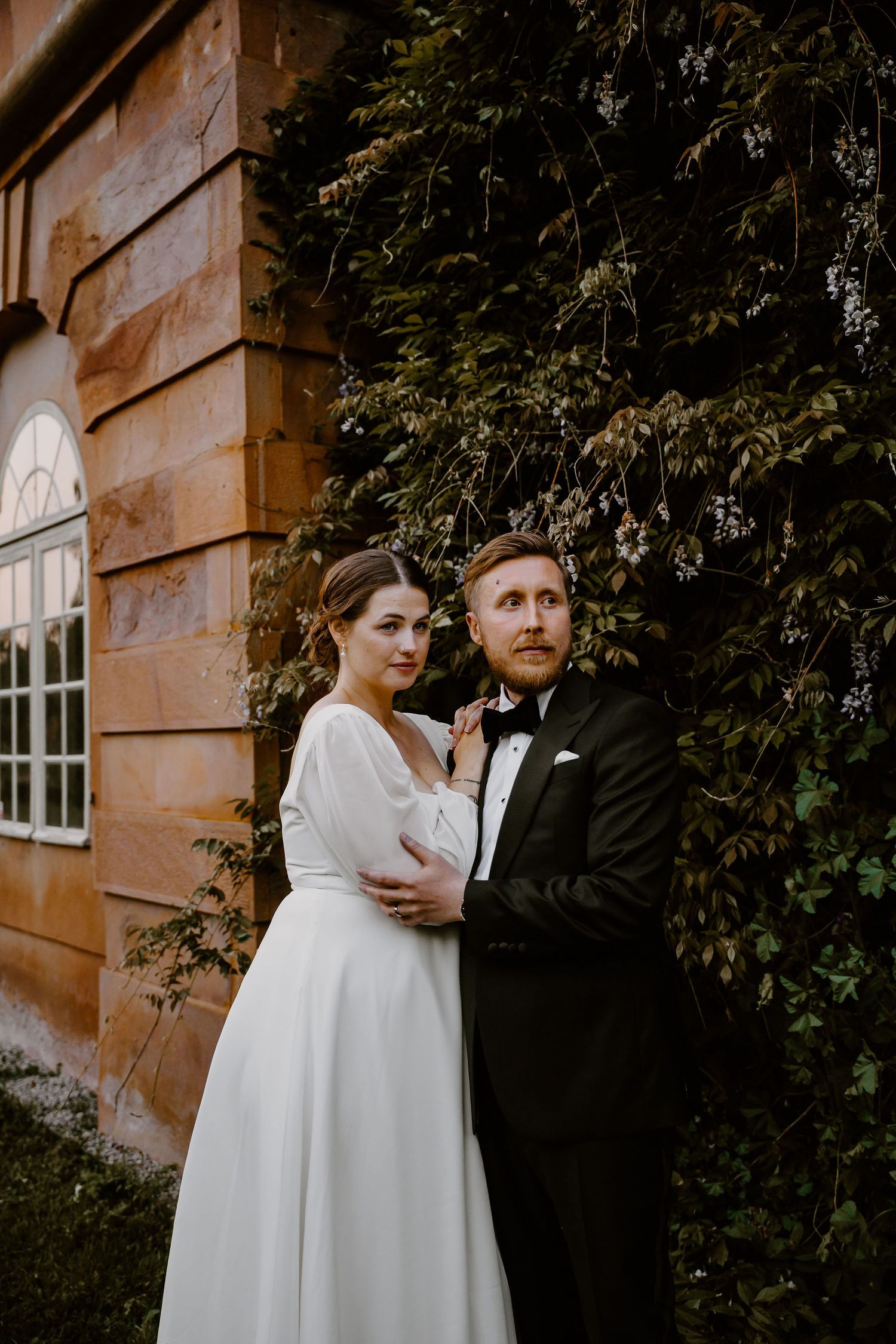 A bride and groom pose together outside a rustic building with ivy-covered walls during their wedding photoshoot.