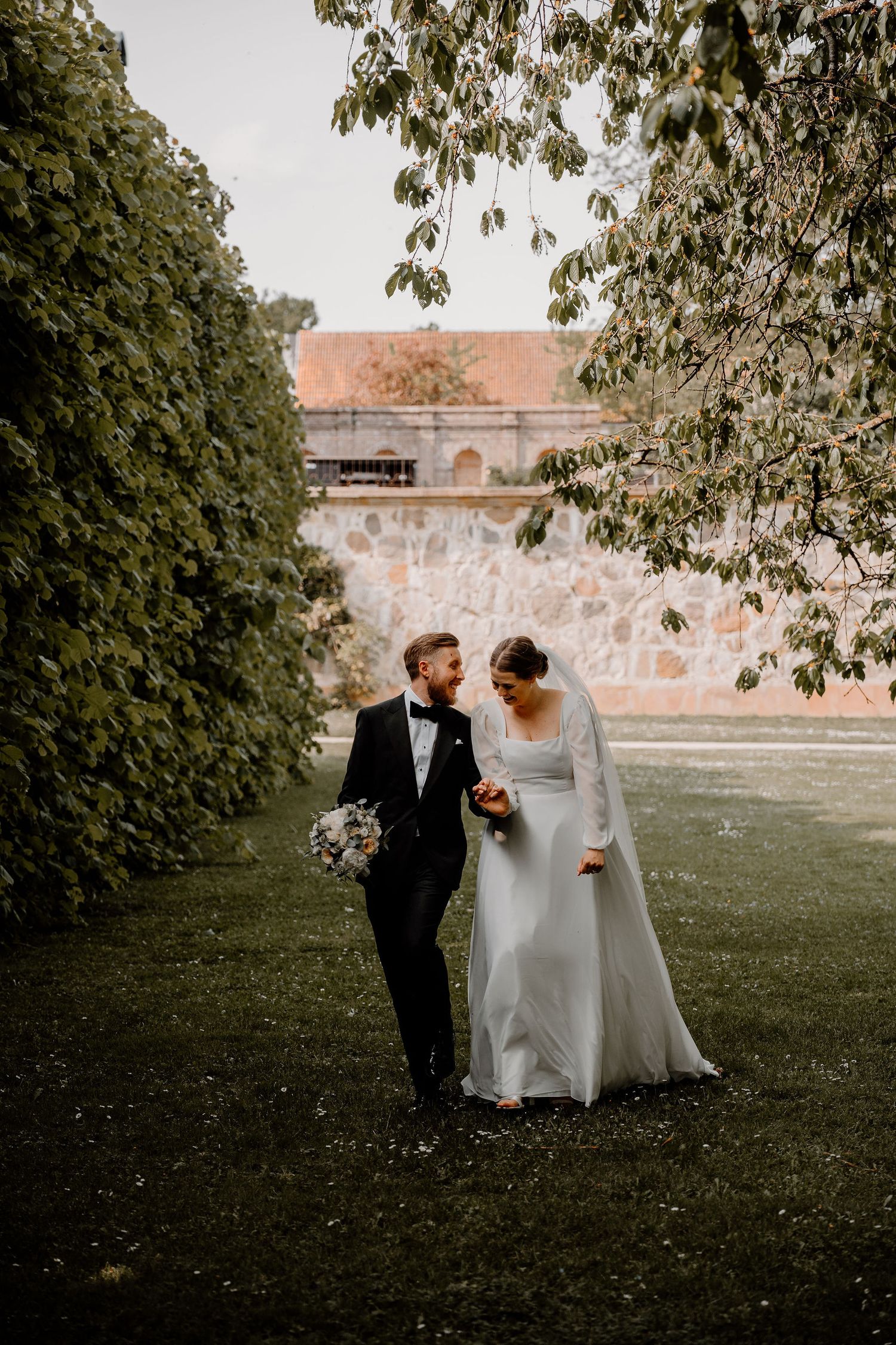 Newlyweds stroll together through a lush garden with historic stone walls and overhanging trees in the background.
