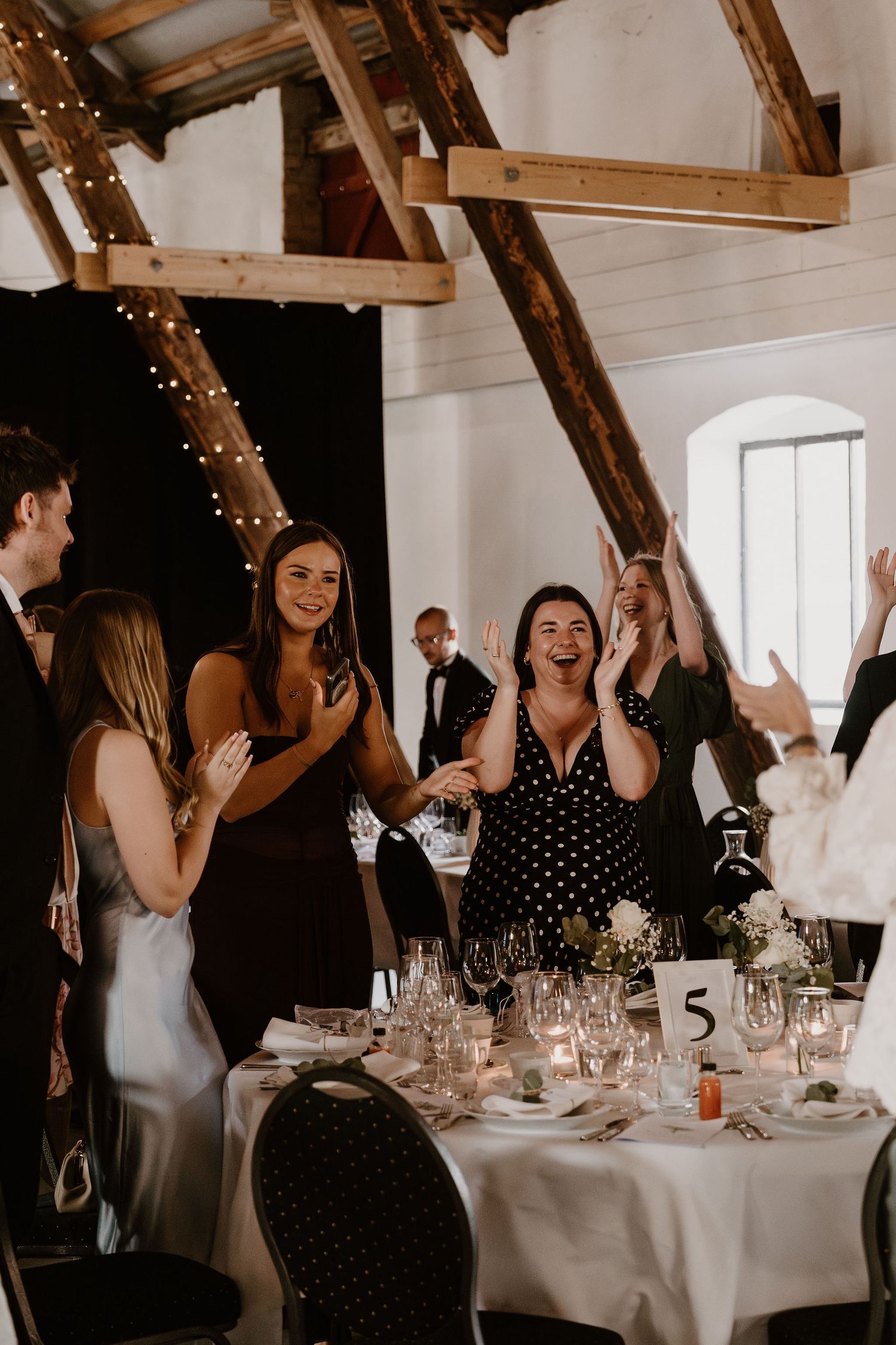 Wedding guests socialize and laugh around elegant white-clothed dining tables under rustic wooden beams with twinkle lights.