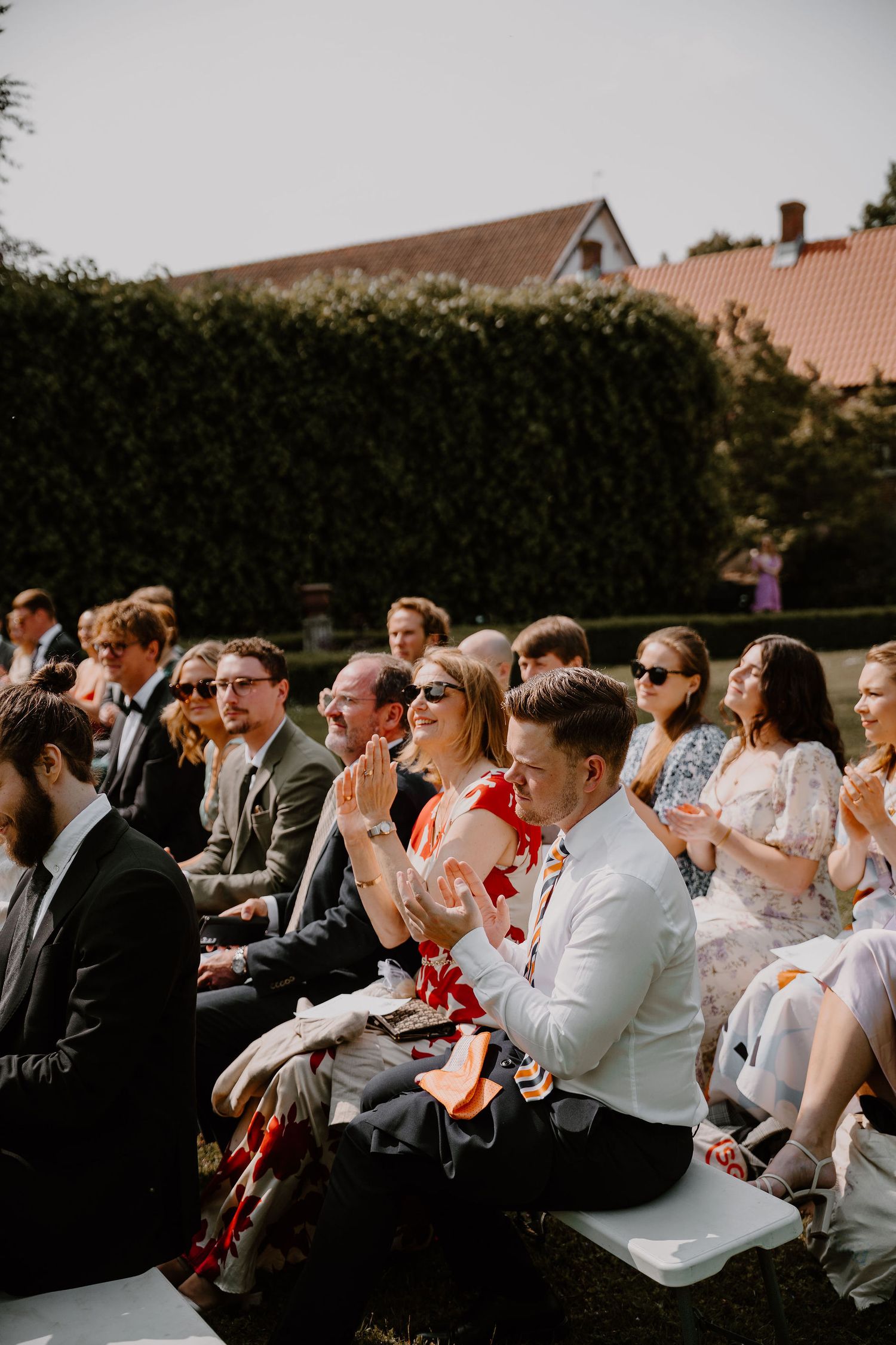 Wedding guests sit on white chairs outdoors watching an emotional ceremony moment unfold.