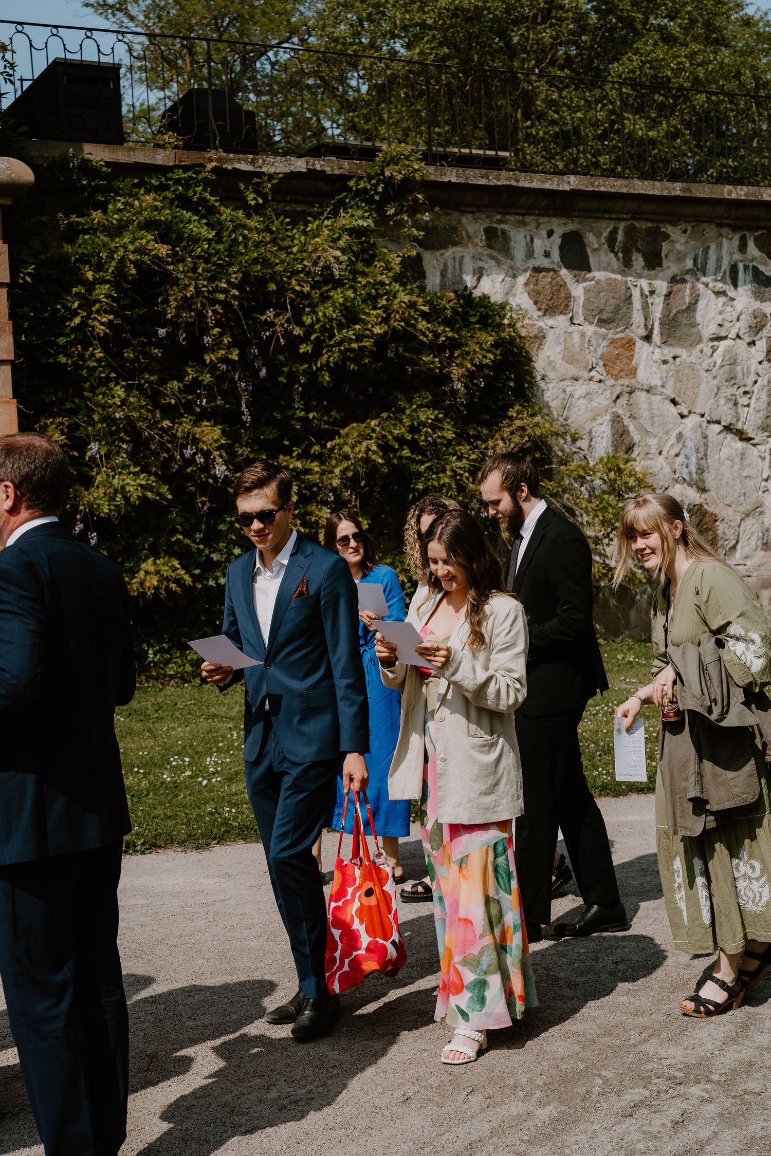 Group of guests walking through a sun-dappled stone wall pathway during an outdoor celebration.