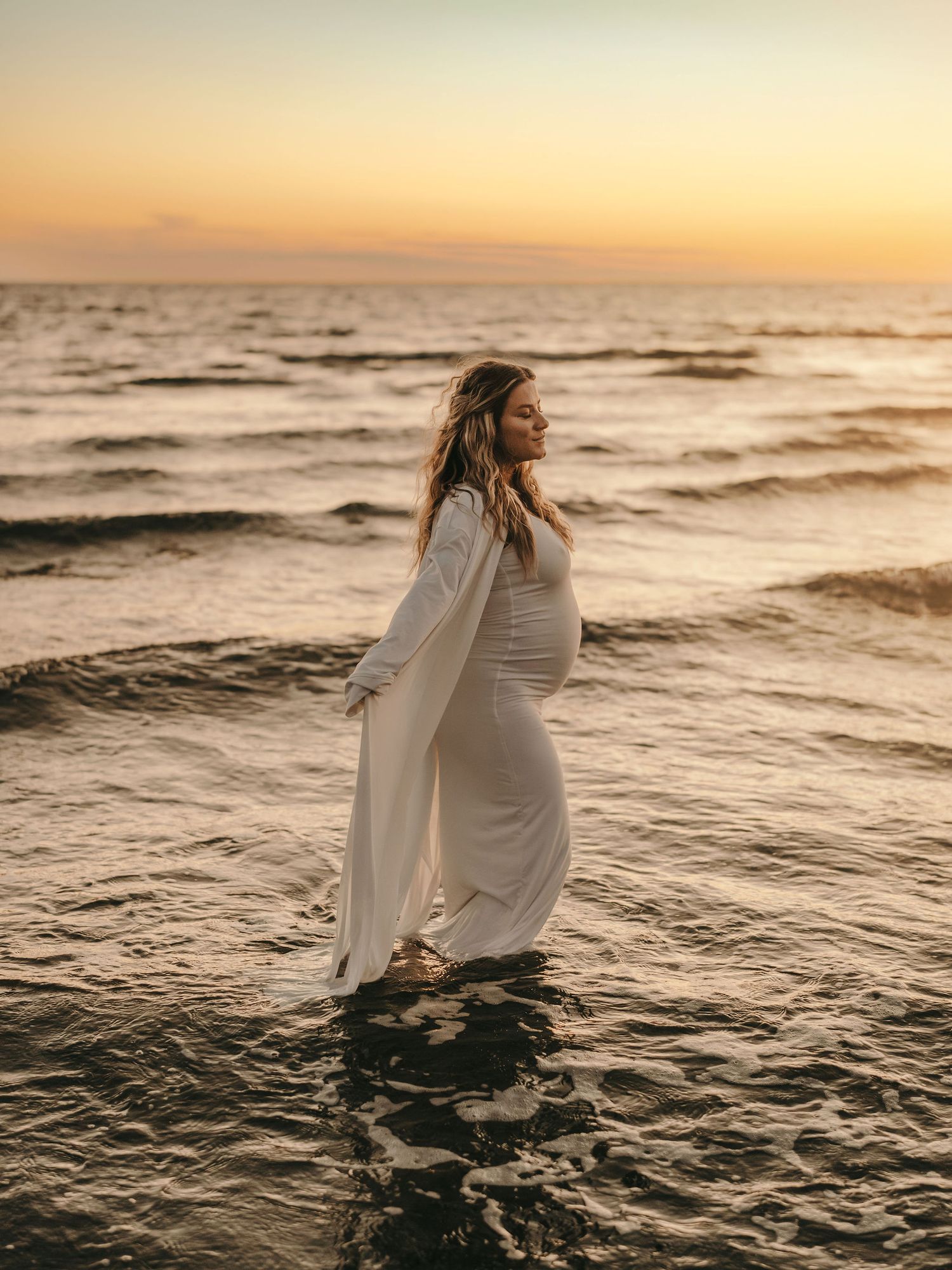 A figure in a flowing white maternity dress stands in ocean waves at sunset, creating a dreamy silhouette against the horizon.