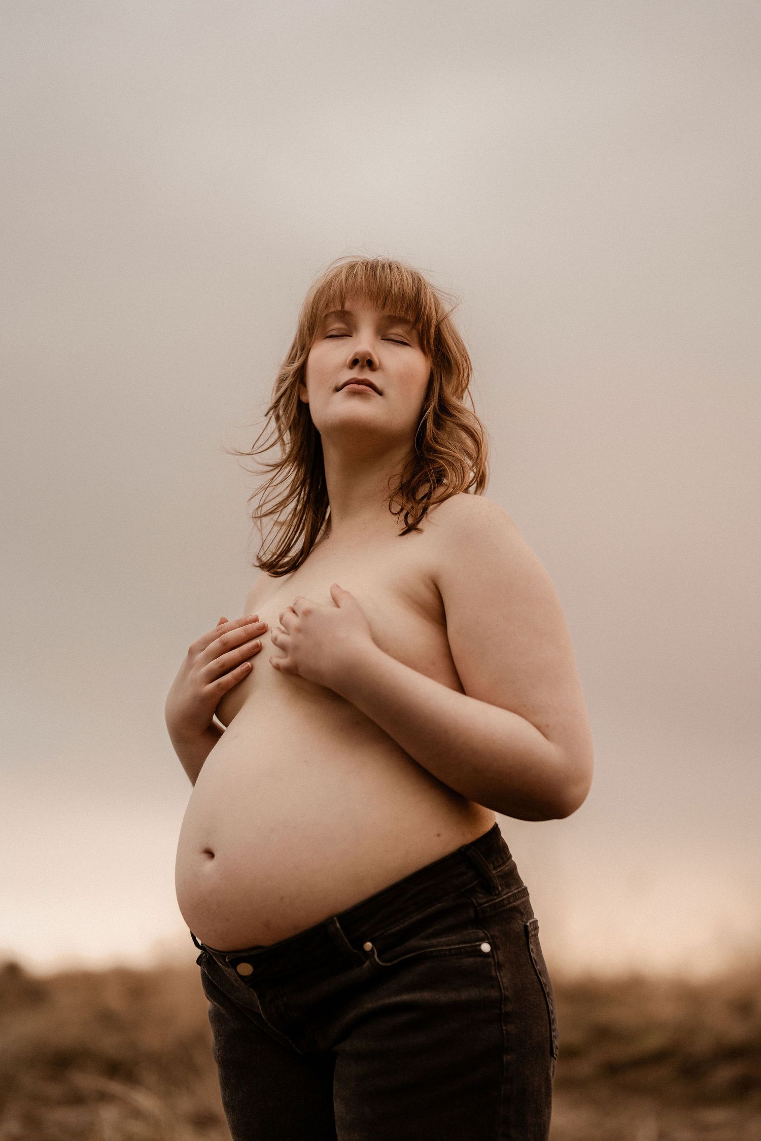 Artistic maternity portrait in sepia tones shows an expectant mother against moody sky background wearing denim.