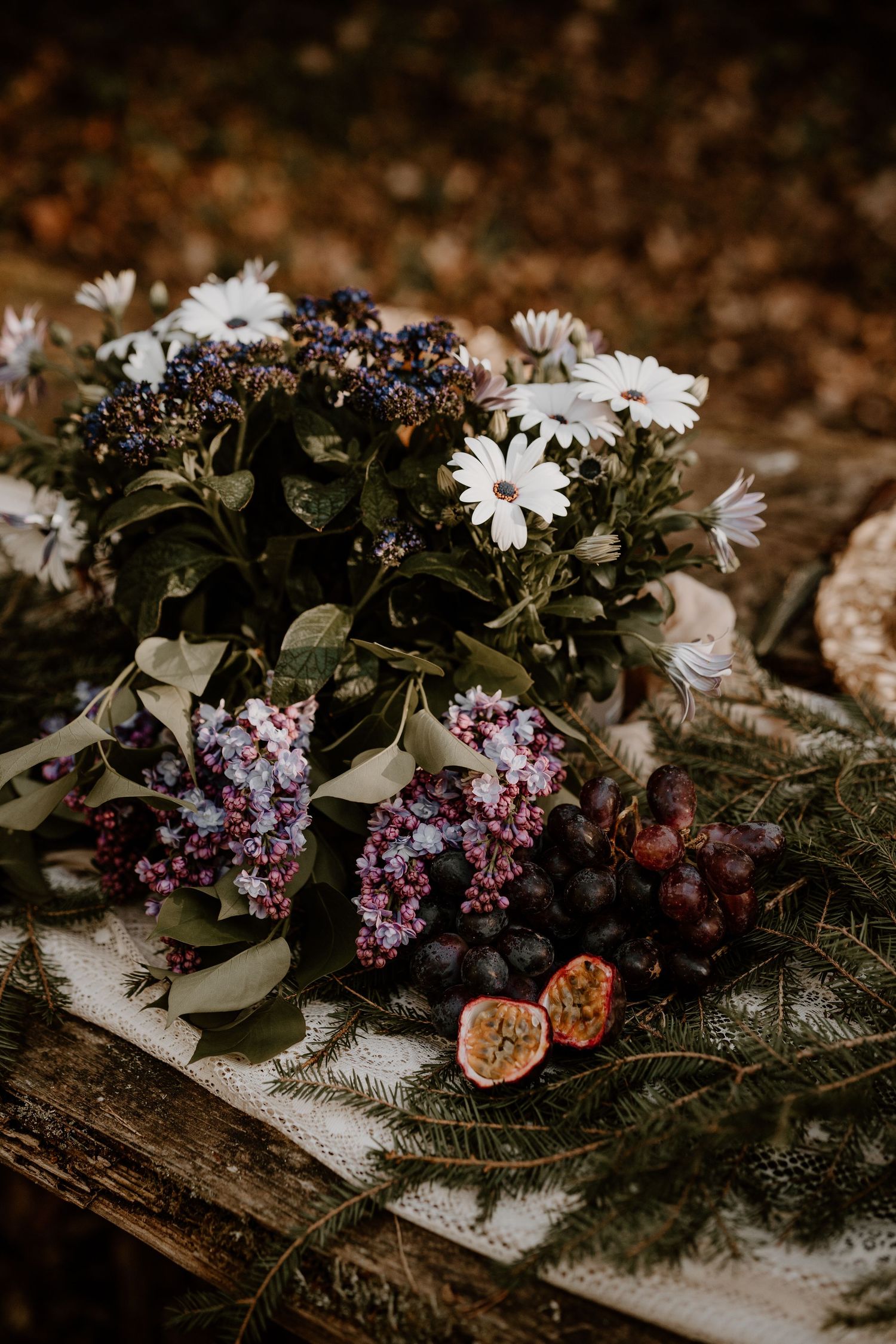 A moody still life arrangement of white daisies, purple lilacs, and cut figs on rustic wood with evergreen sprigs.