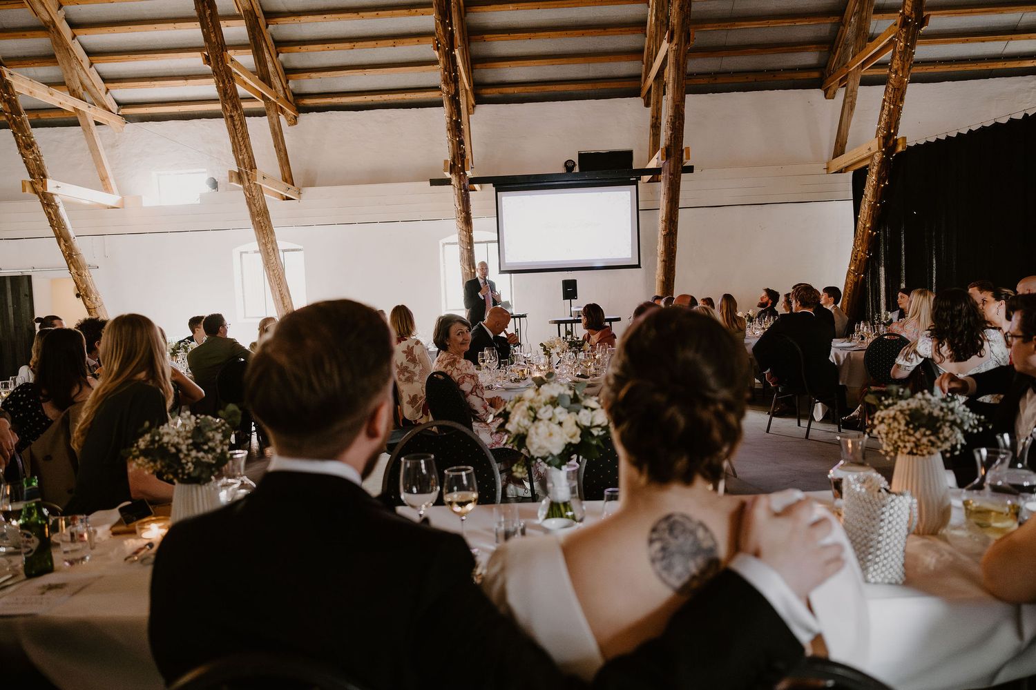 Wedding guests seated at decorated reception tables inside rustic barn venue with wood beams and white walls.
