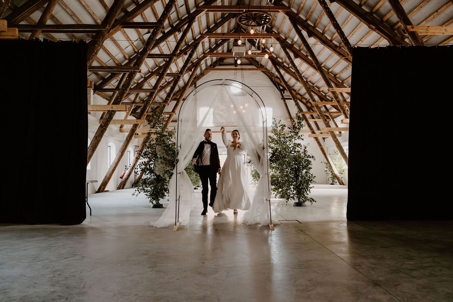 A bride and groom walk together under a rustic wooden beam ceiling decorated with minimal greenery in an A-frame venue.