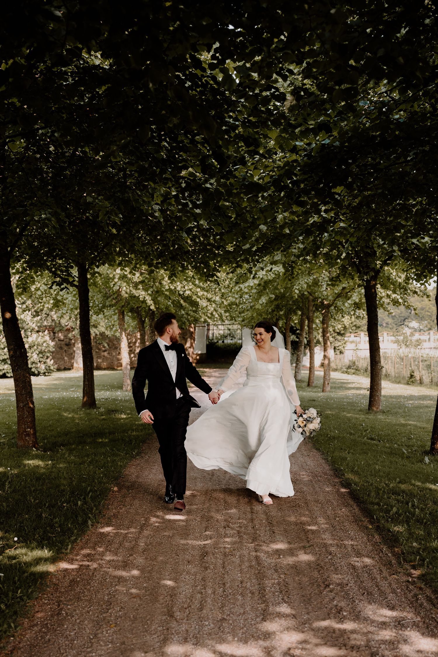 Newlyweds walk playfully together down a scenic tree-lined dirt path during their wedding photoshoot.