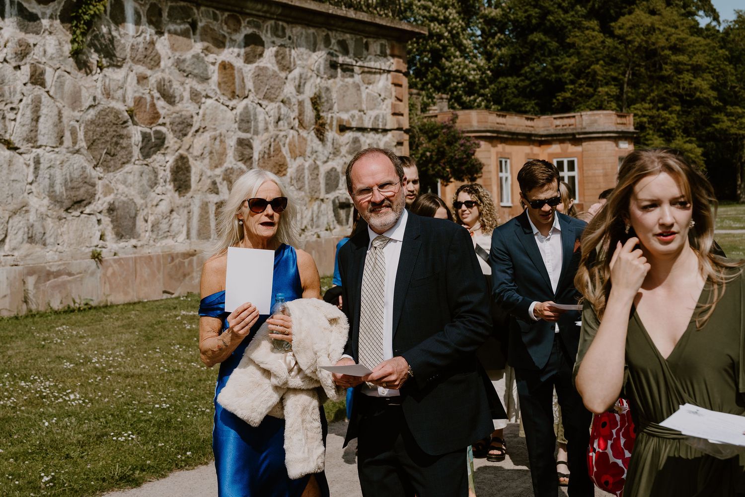 Guests gather outdoors on a lawn near a stone wall during a social event on a sunny day.