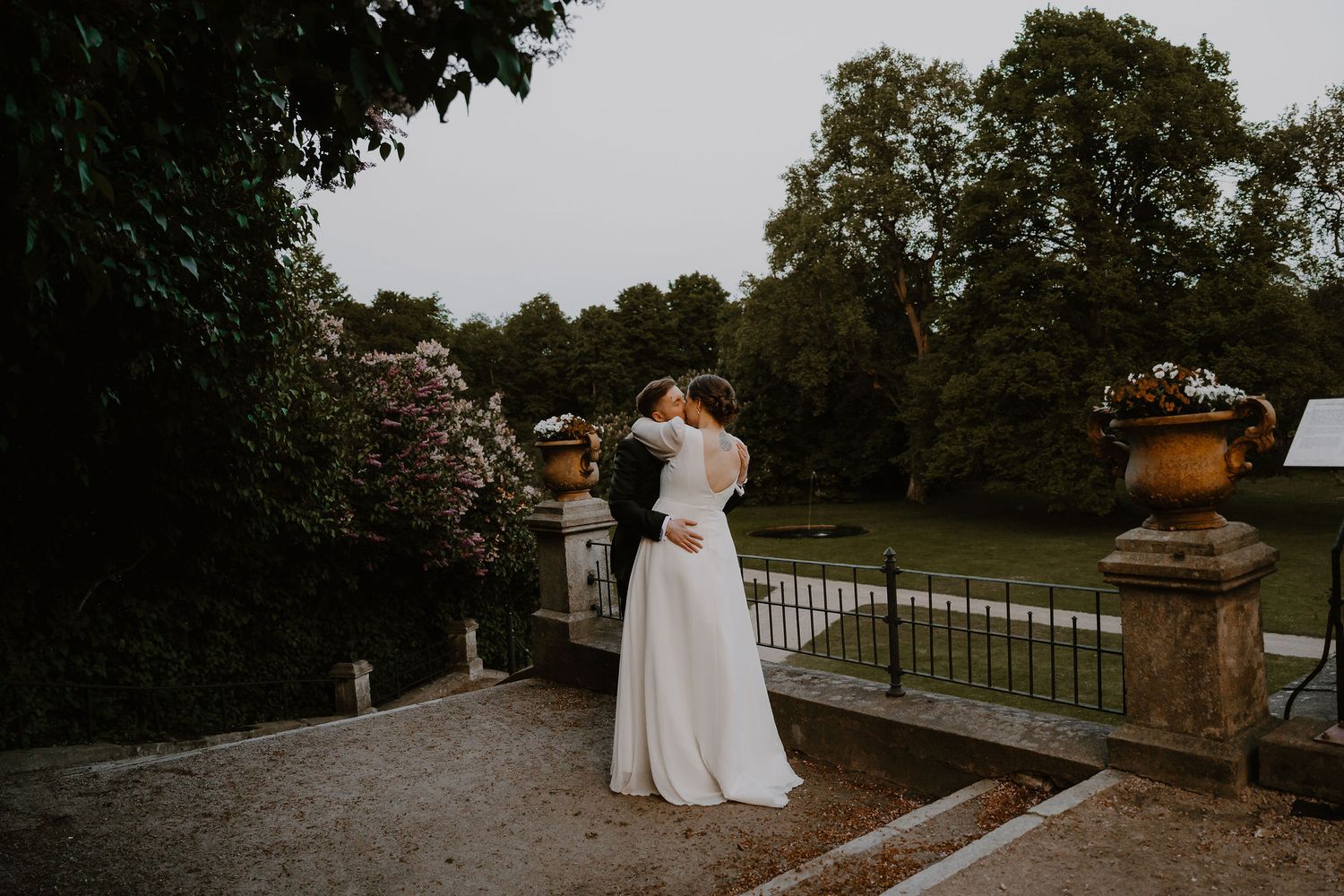 A couple embraces in formal wedding attire on a stone terrace overlooking scenic grounds at dusk.