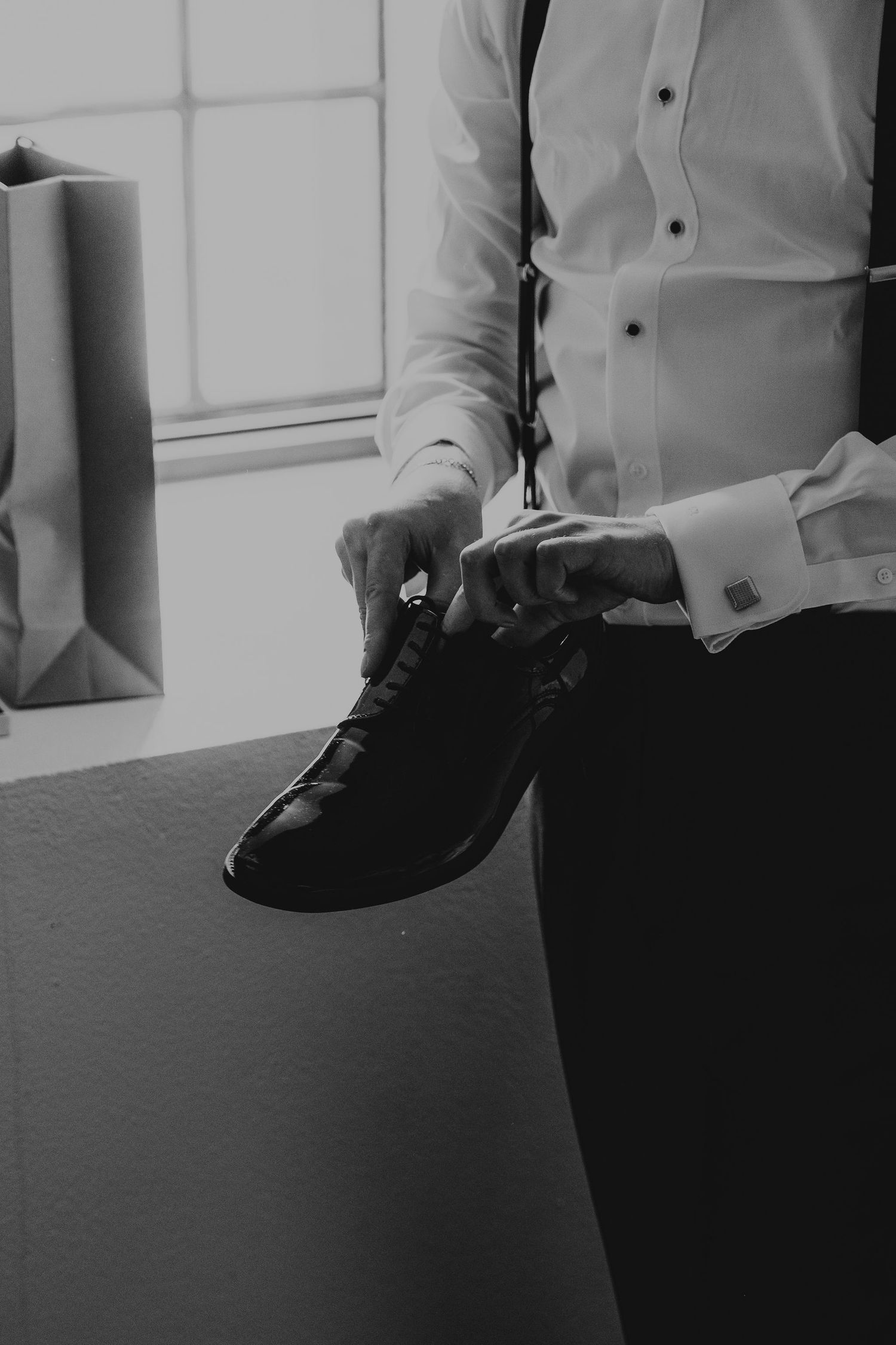 Black and white photo of someone tying formal dress shoes while wearing a crisp white button-down shirt.
