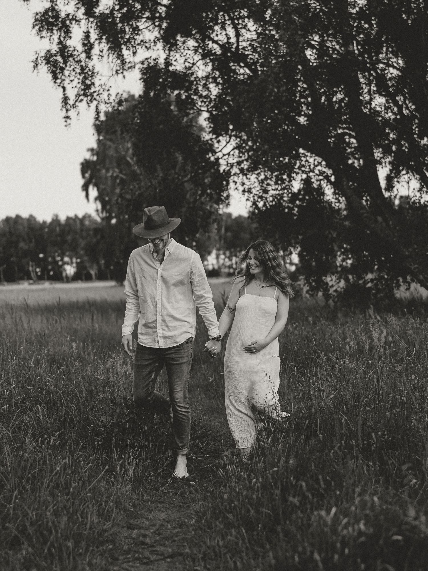 A couple walking hand in hand through a grassy field under trees in black and white vintage photography.