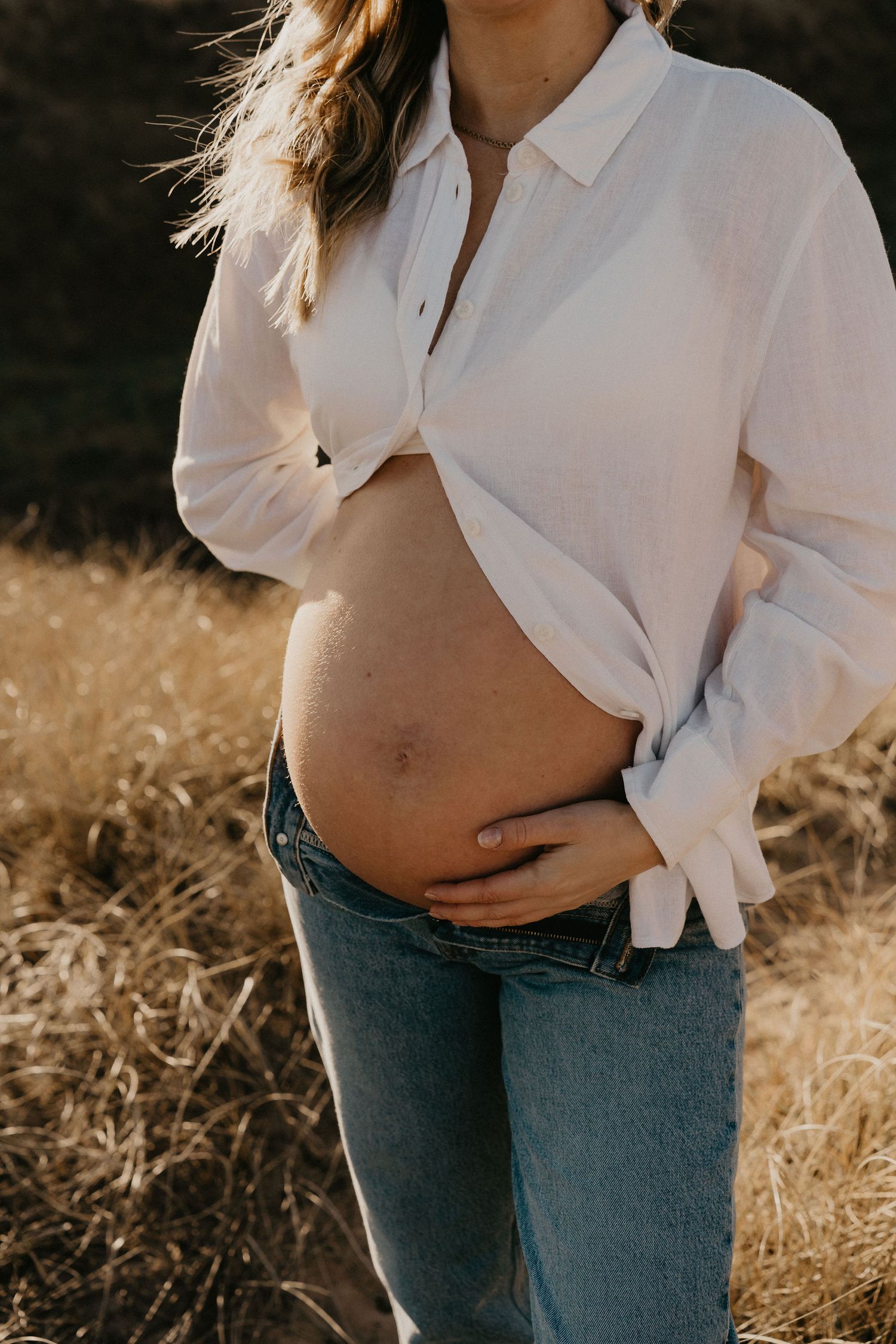 A pregnant woman in a white button-down shirt and jeans cradles her bare belly in golden sunlit field.