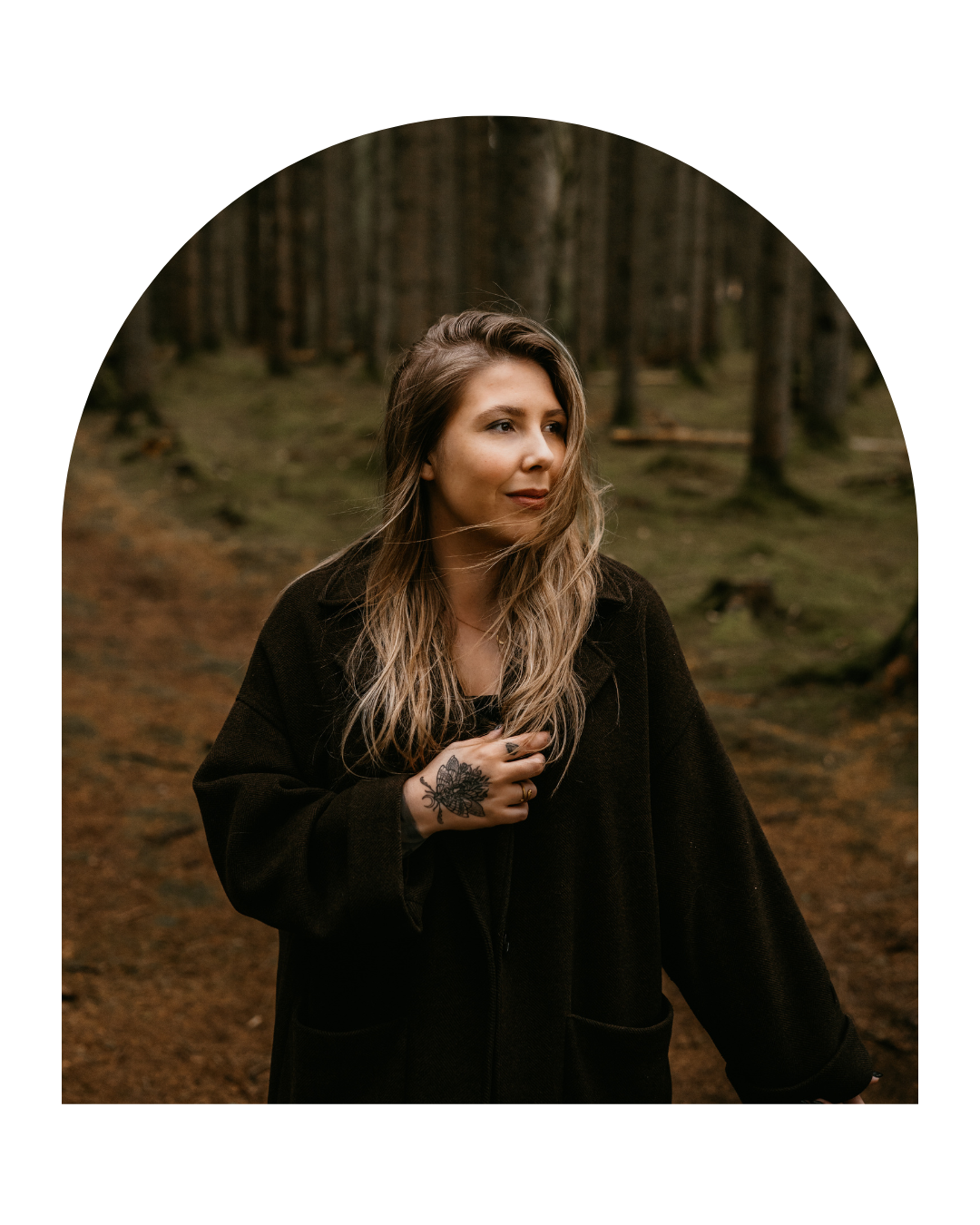 Person in dark clothing poses thoughtfully in moody forest setting with brown leaves on winding path.