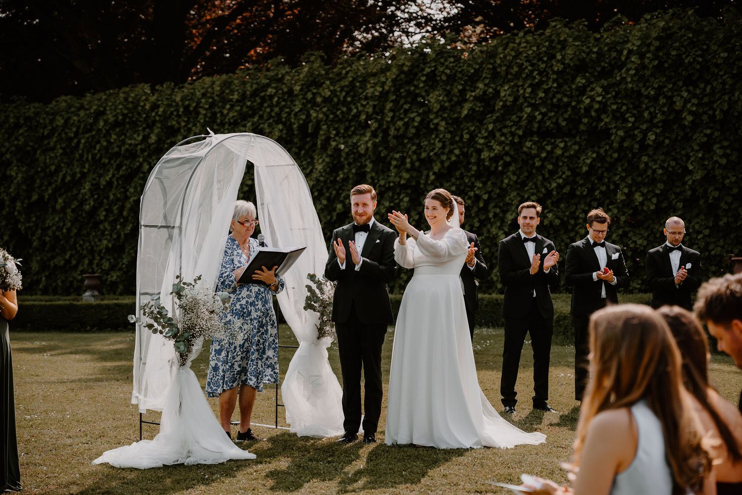 A wedding ceremony takes place outdoors with a white arch backdrop and guests on a green lawn.