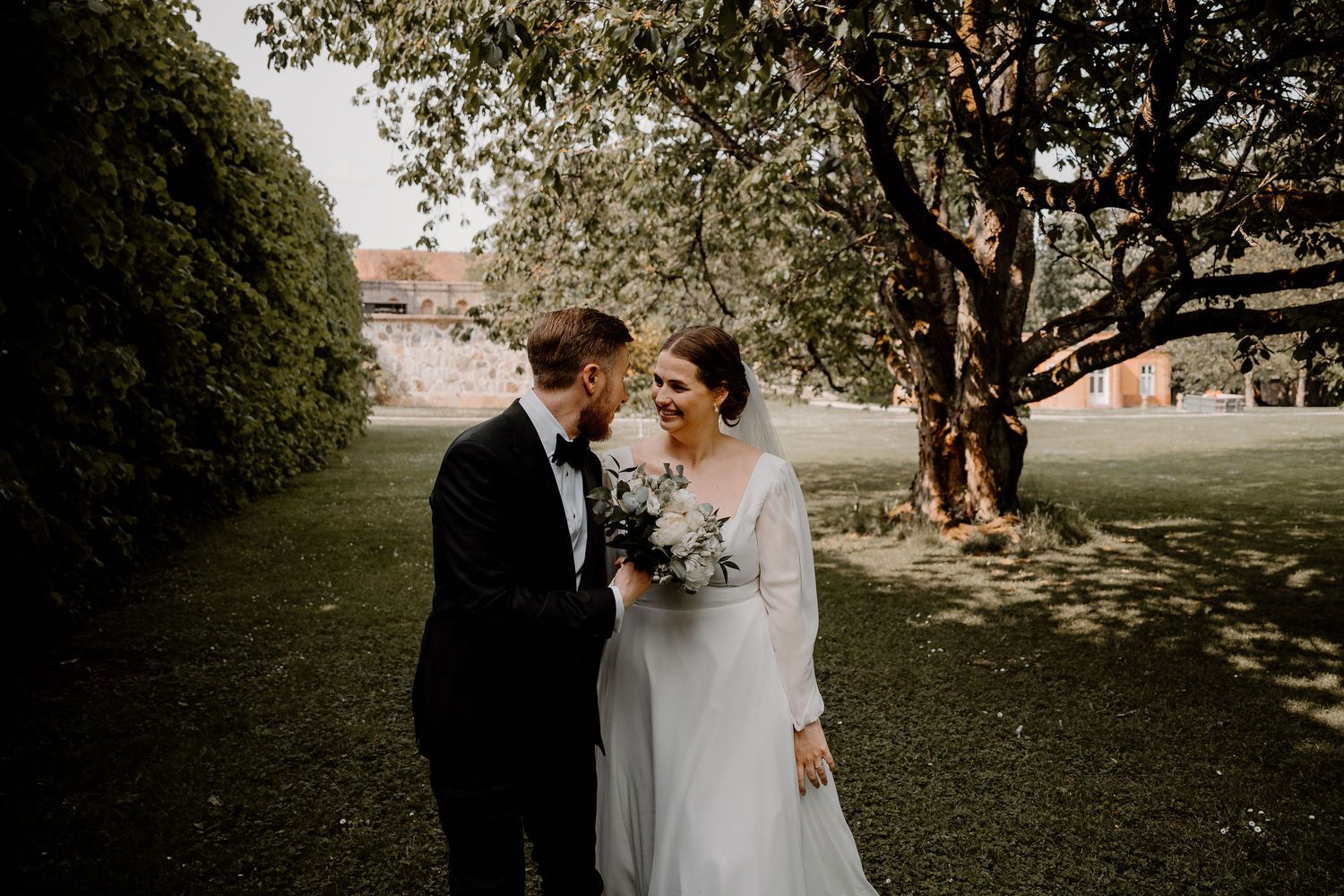 A couple in formal wedding attire stands close together beneath trees on a grassy lawn.