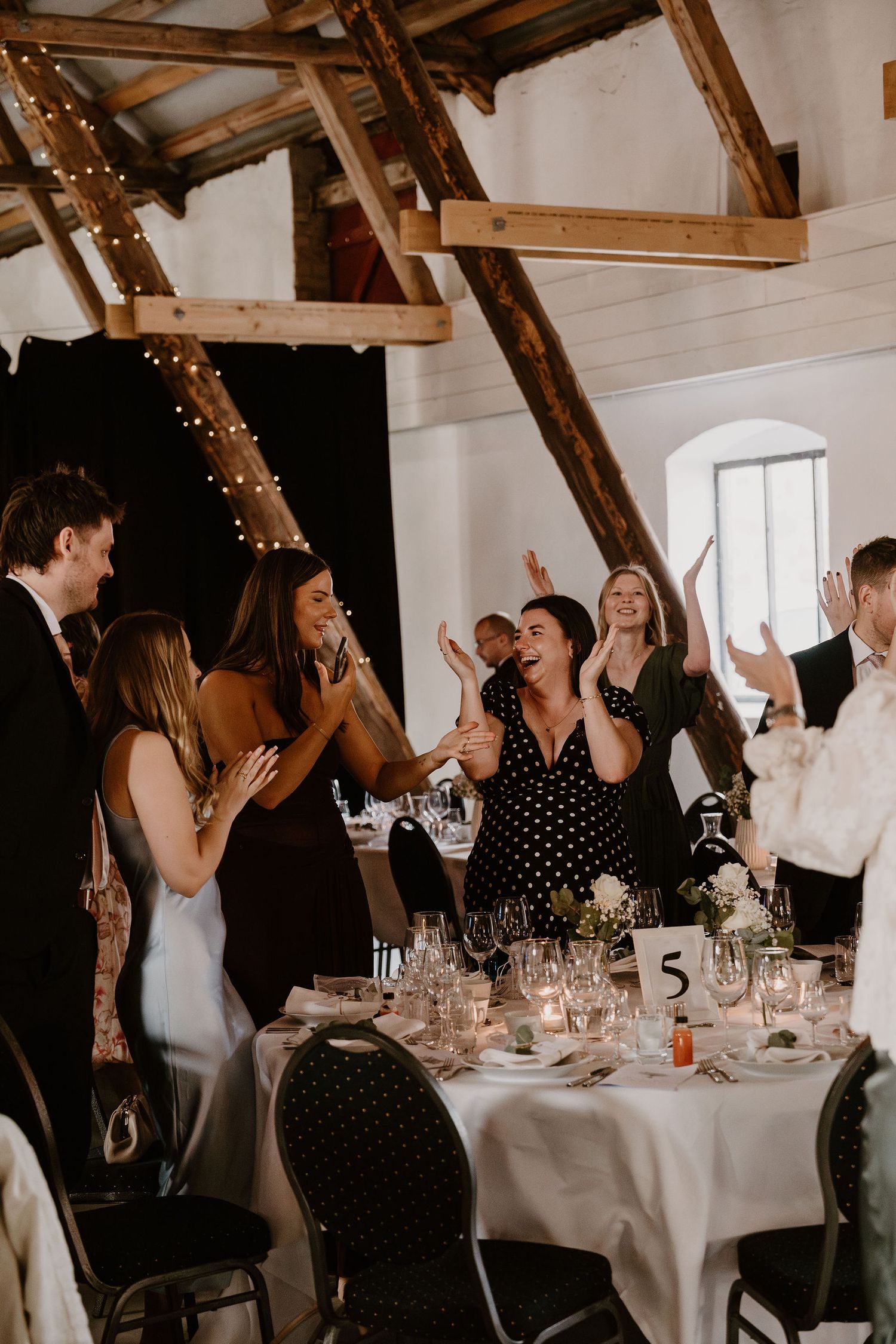 Wedding guests in formal attire gather around elegant round tables with white linens in a rustic barn venue with string lights.