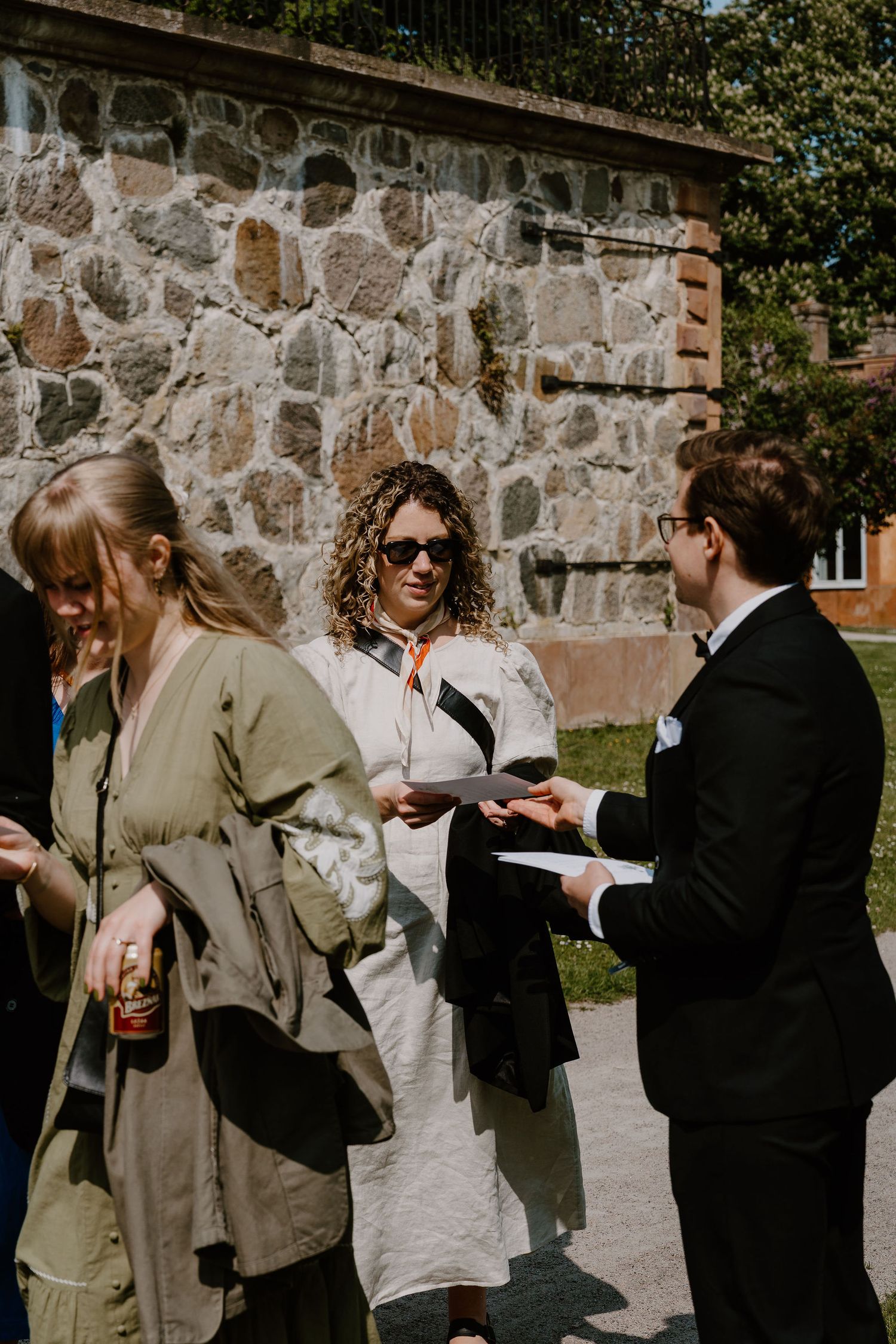 People gathered at an outdoor event chatting near a stone wall on a sunny day.