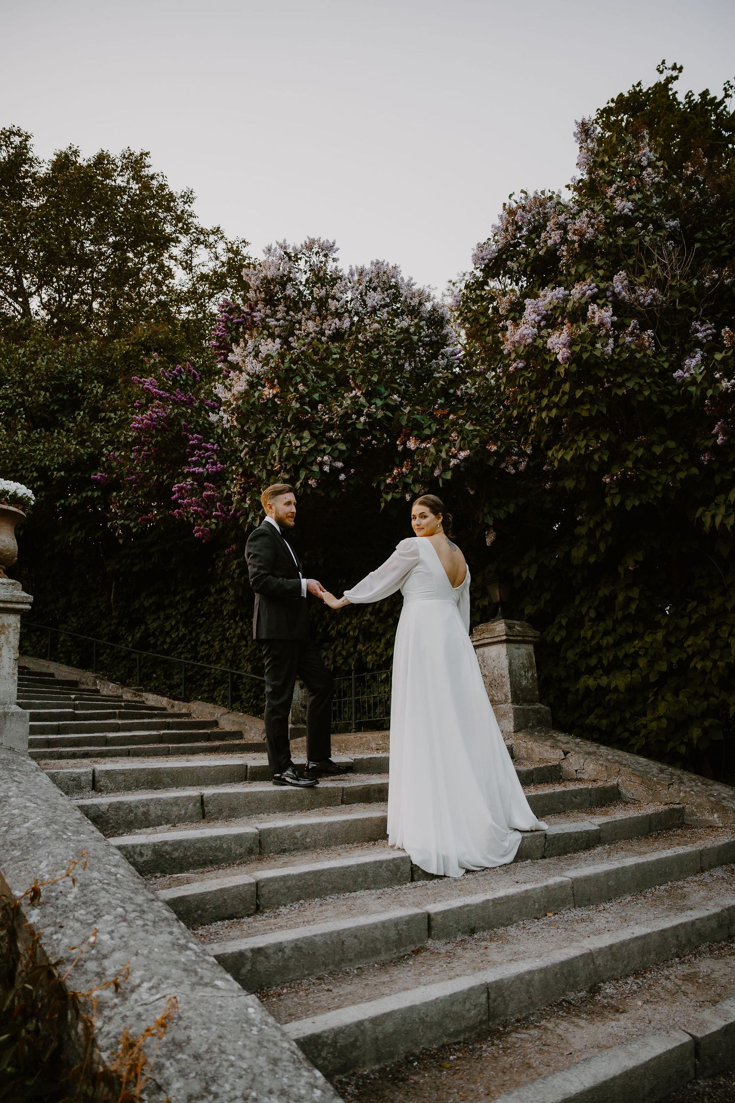 A couple walks up stone steps surrounded by lush greenery in a romantic garden setting during a wedding photo session.