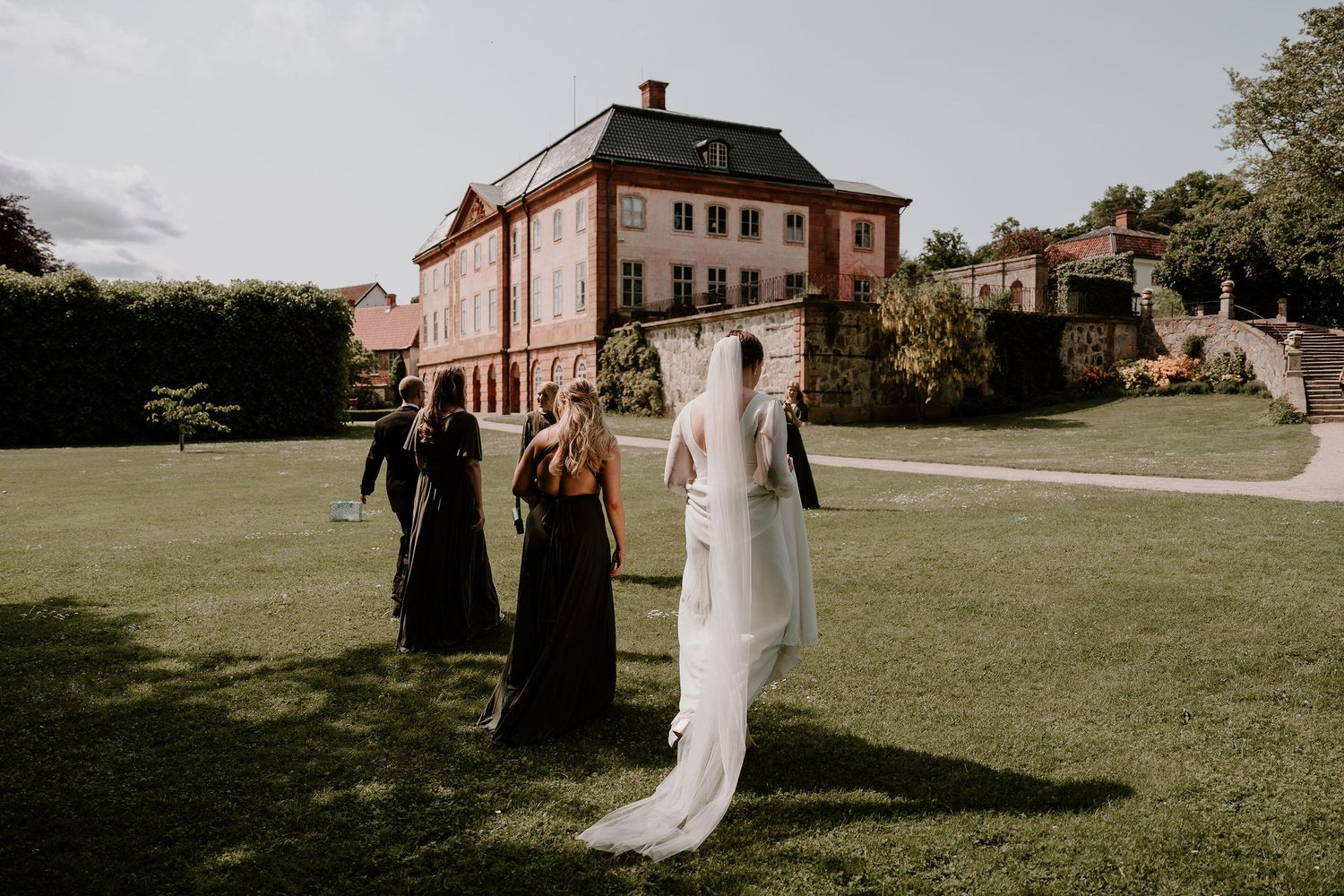A wedding party walks across a manicured lawn in front of a grand historic mansion on a summer evening.