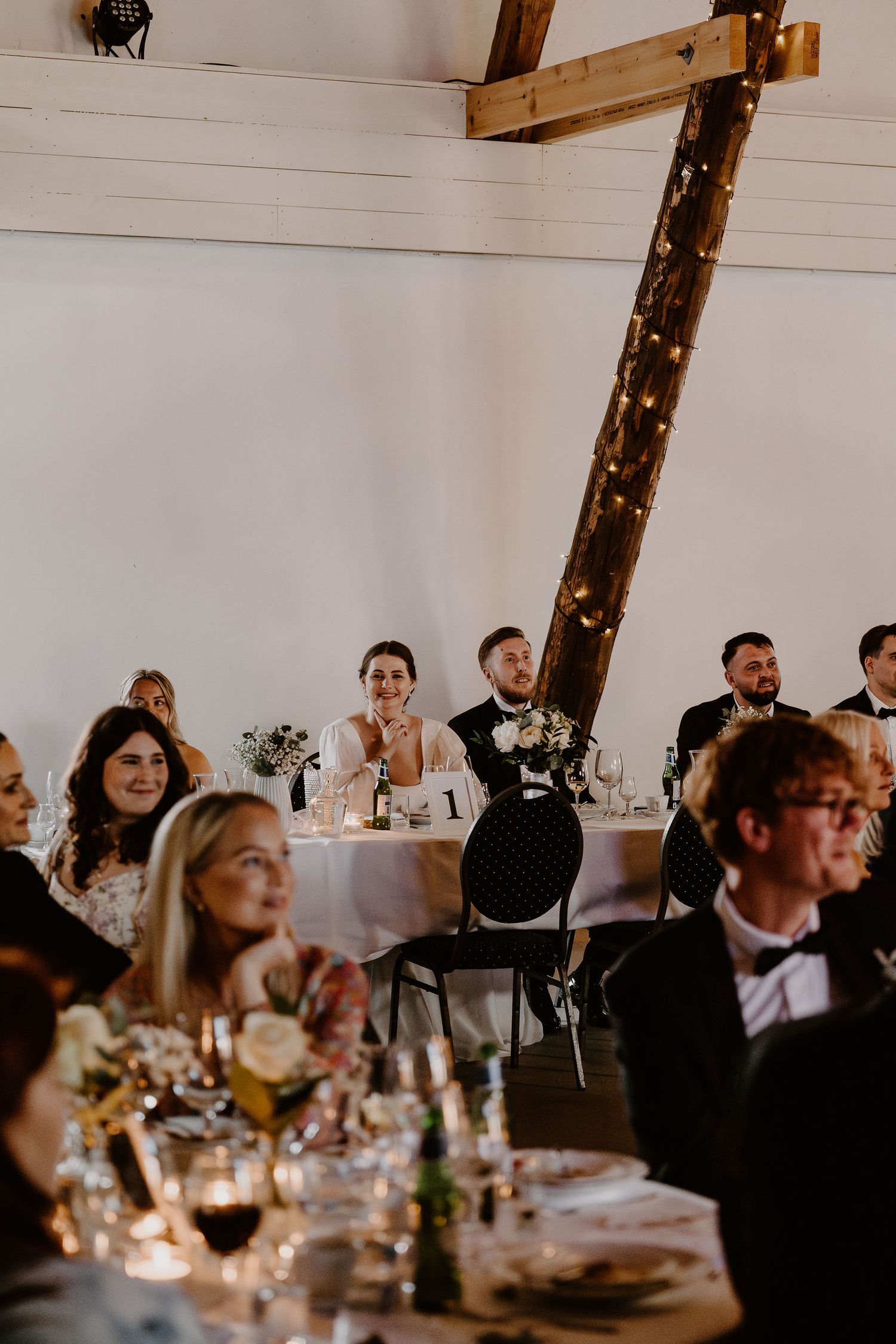 Guests gathered at elegantly set dinner tables with wine glasses and candlelight in a rustic barn venue.