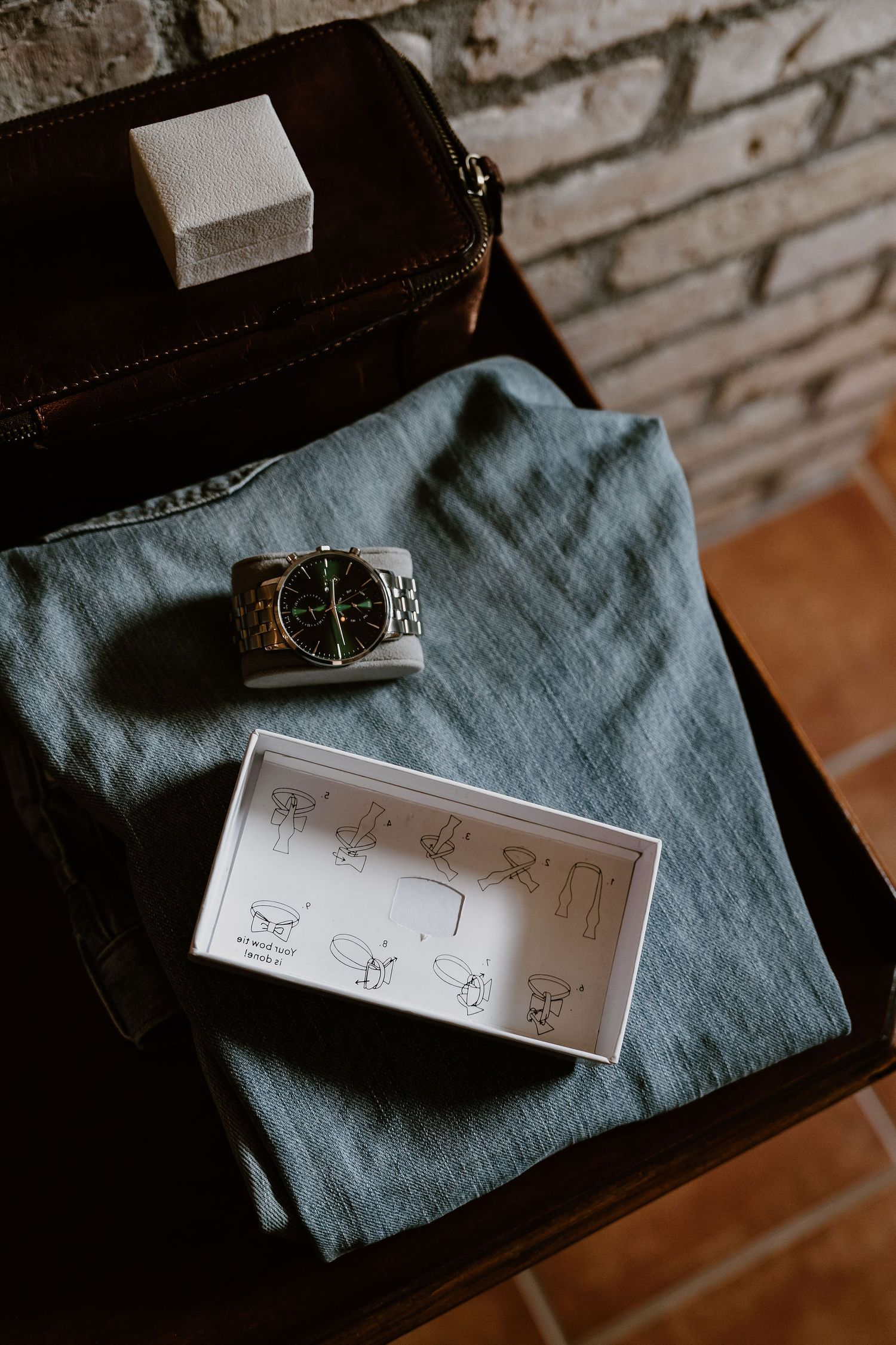 Wedding accessories including a watch and illustrated cufflinks guide arranged on blue fabric against brick wall.