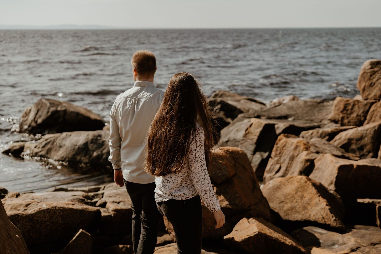Couple stands together on rocky coastal shoreline overlooking misty ocean during golden hour.