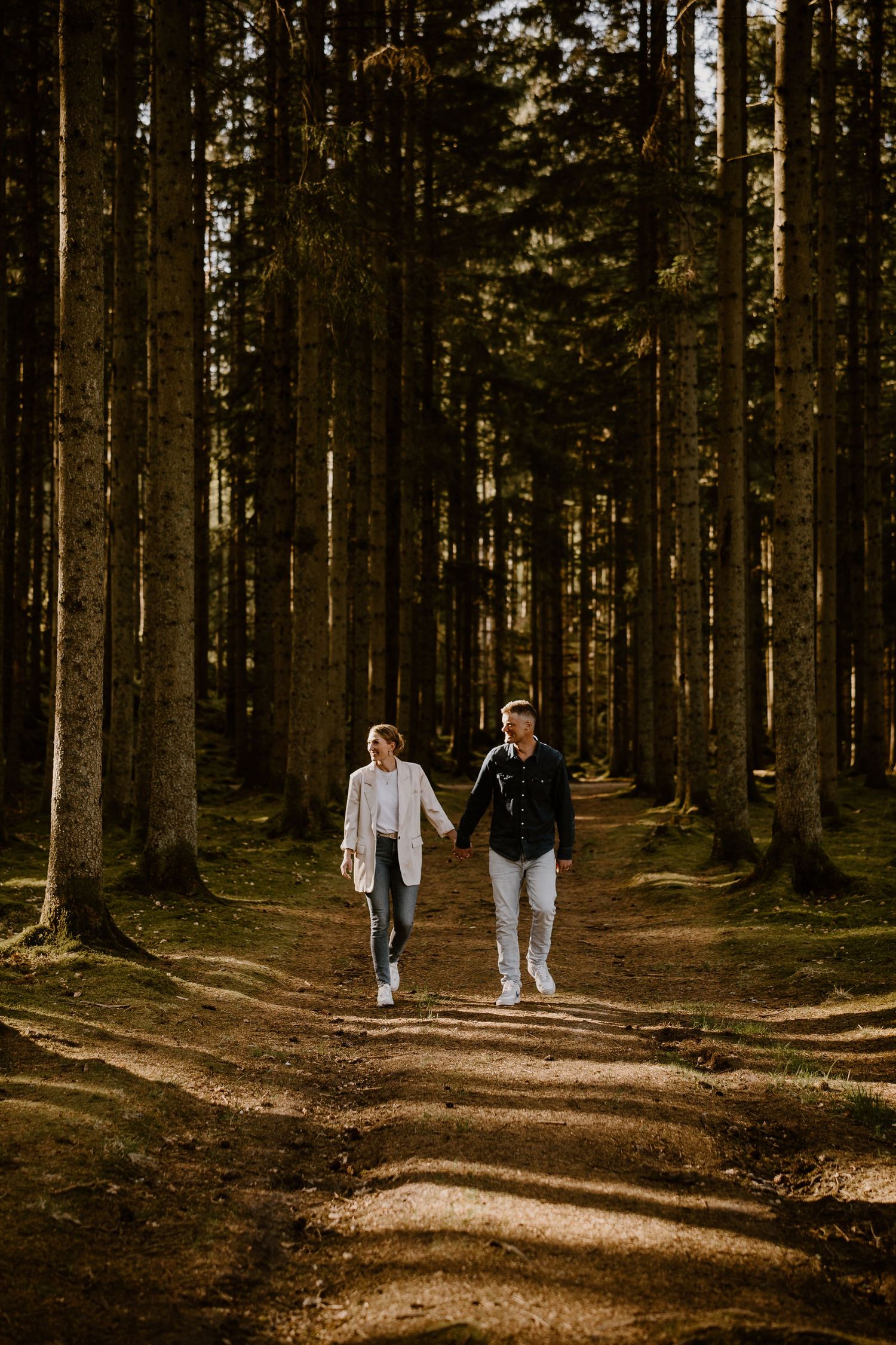 Two people walk together along a sunlit forest trail surrounded by tall evergreen trees at golden hour.