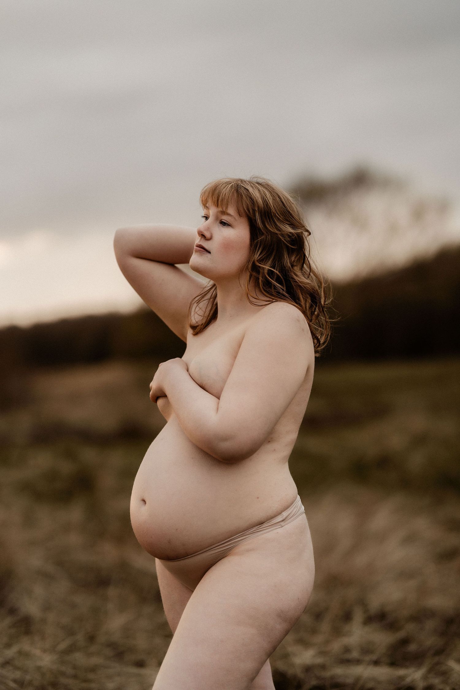 Artistic nude photograph showing a curvy figure posing against a moody natural landscape at dusk.