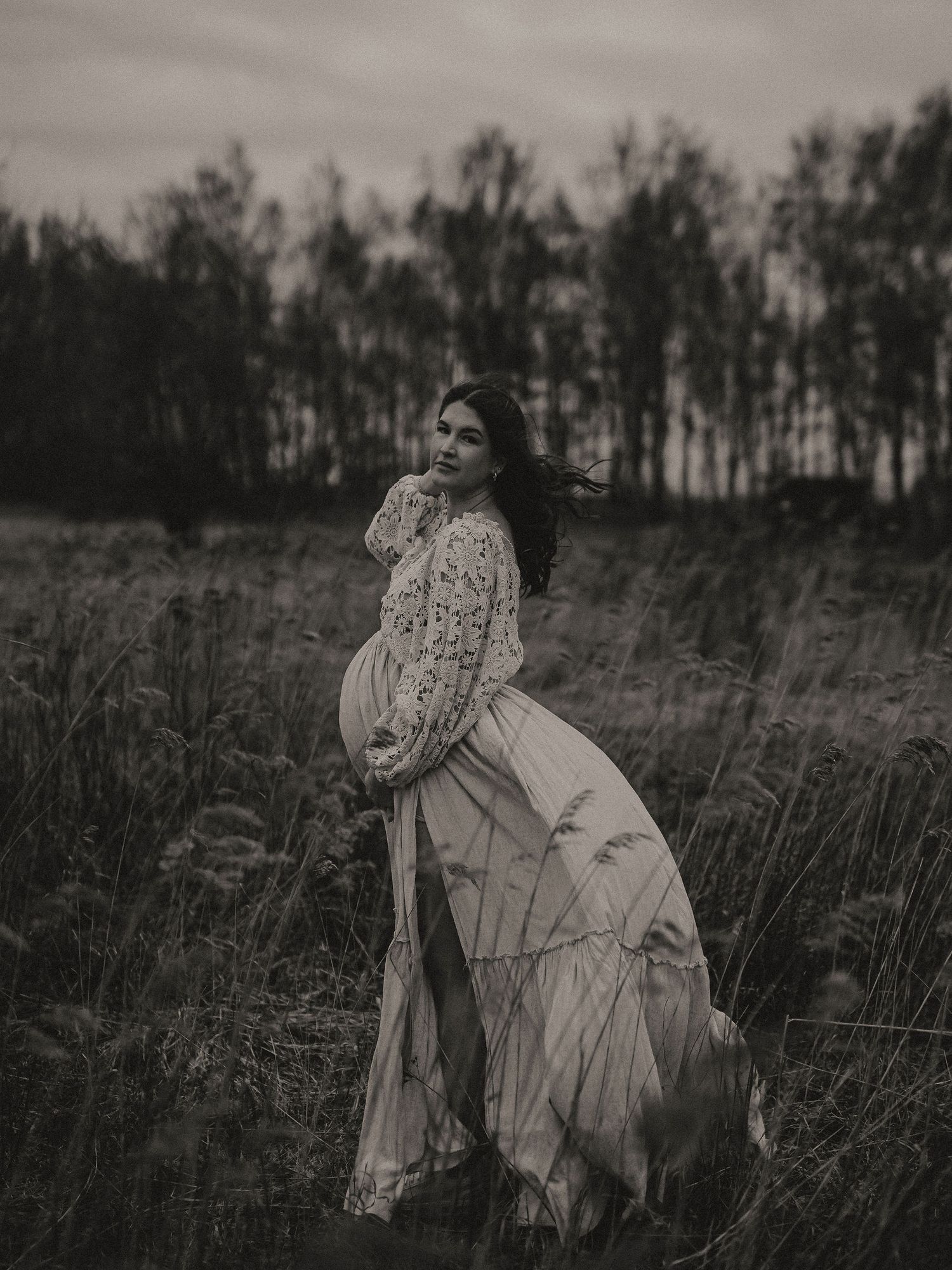 A moody black and white portrait of a person in a flowing white dress standing in a field with trees in the background.