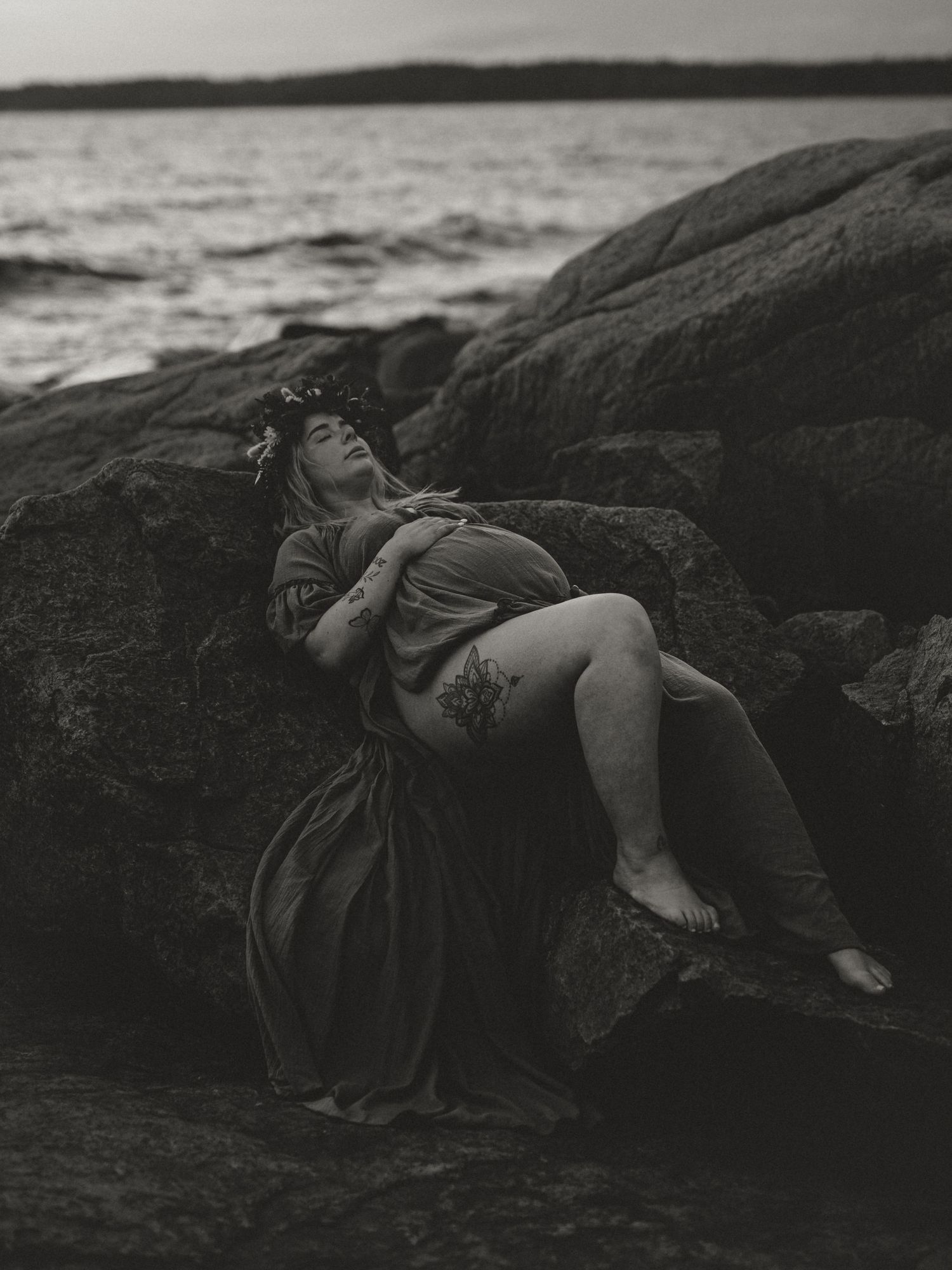 A figure in dark clothing reclines on coastal rocks by the water in a dreamy black and white photograph.