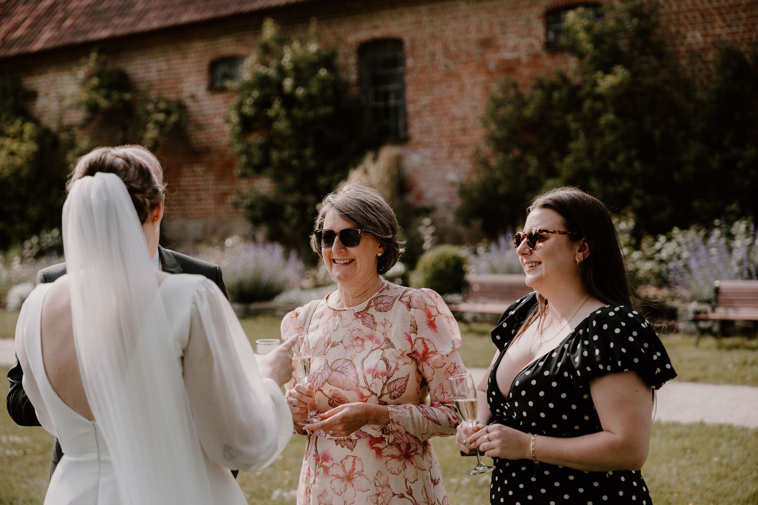 A group of people gather outdoors on a lawn near a brick building wearing formal attire for a wedding celebration.