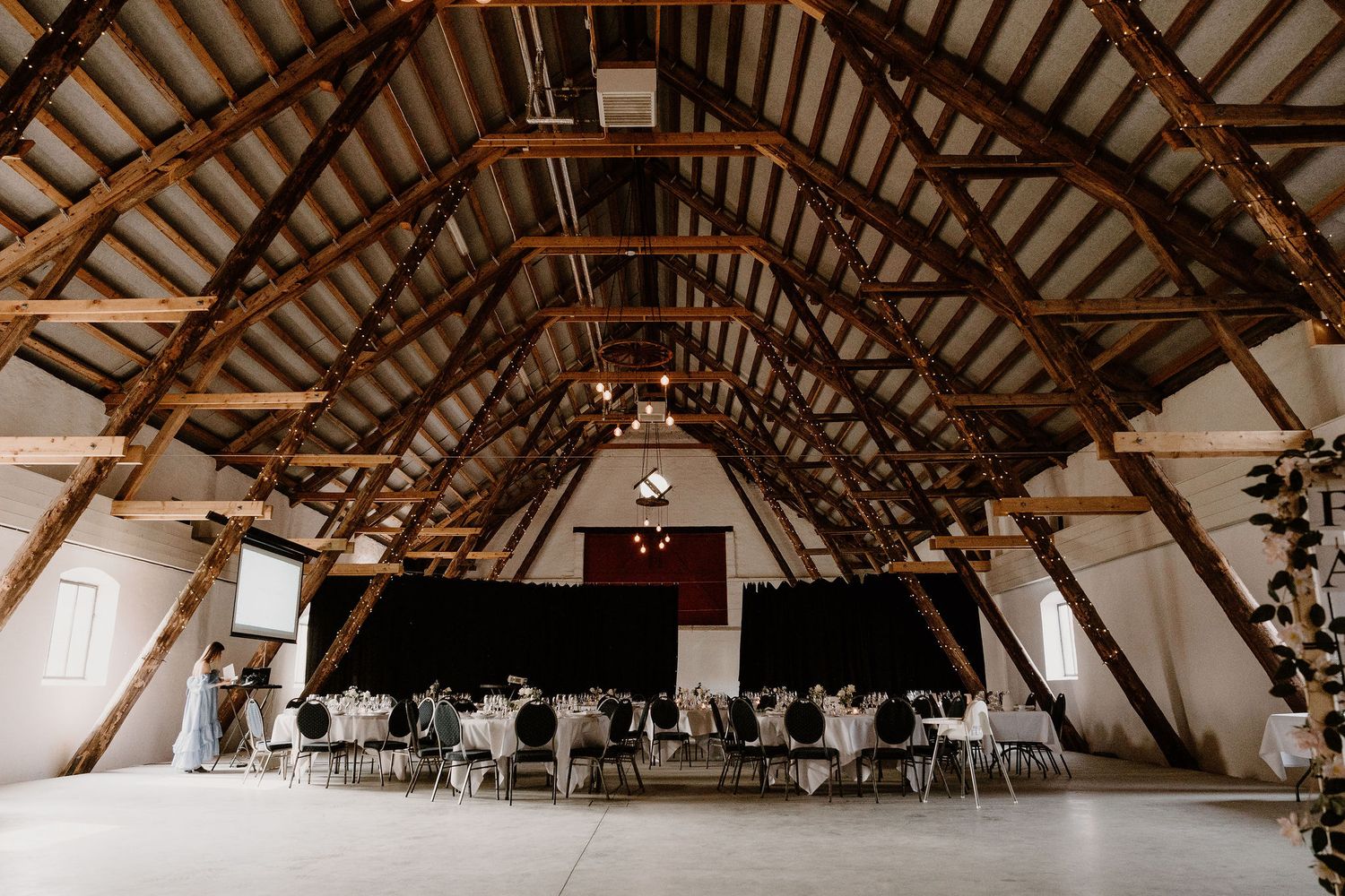 Expansive rustic wooden barn venue interior with exposed beams and white table settings for an elegant wedding reception.