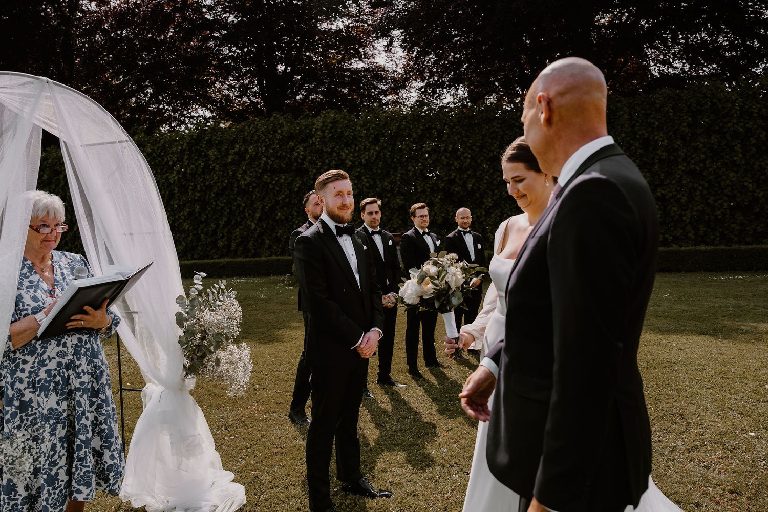 Wedding party gathers outdoors on grassy lawn during outdoor ceremony beneath trees.